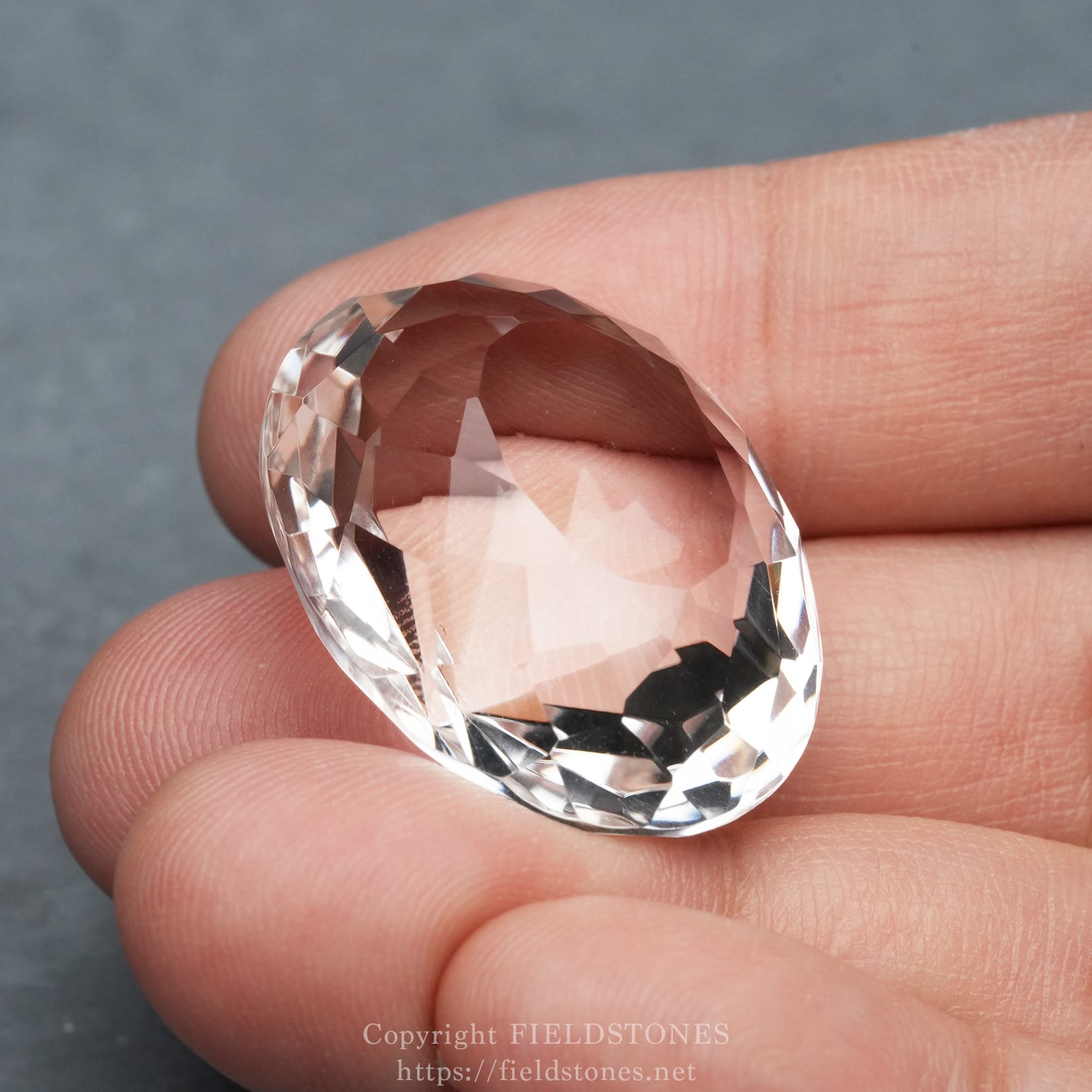 6537-3 - FieldStones Close-up of an oval-cut, faceted clear gemstone held between fingertips against a gray background.