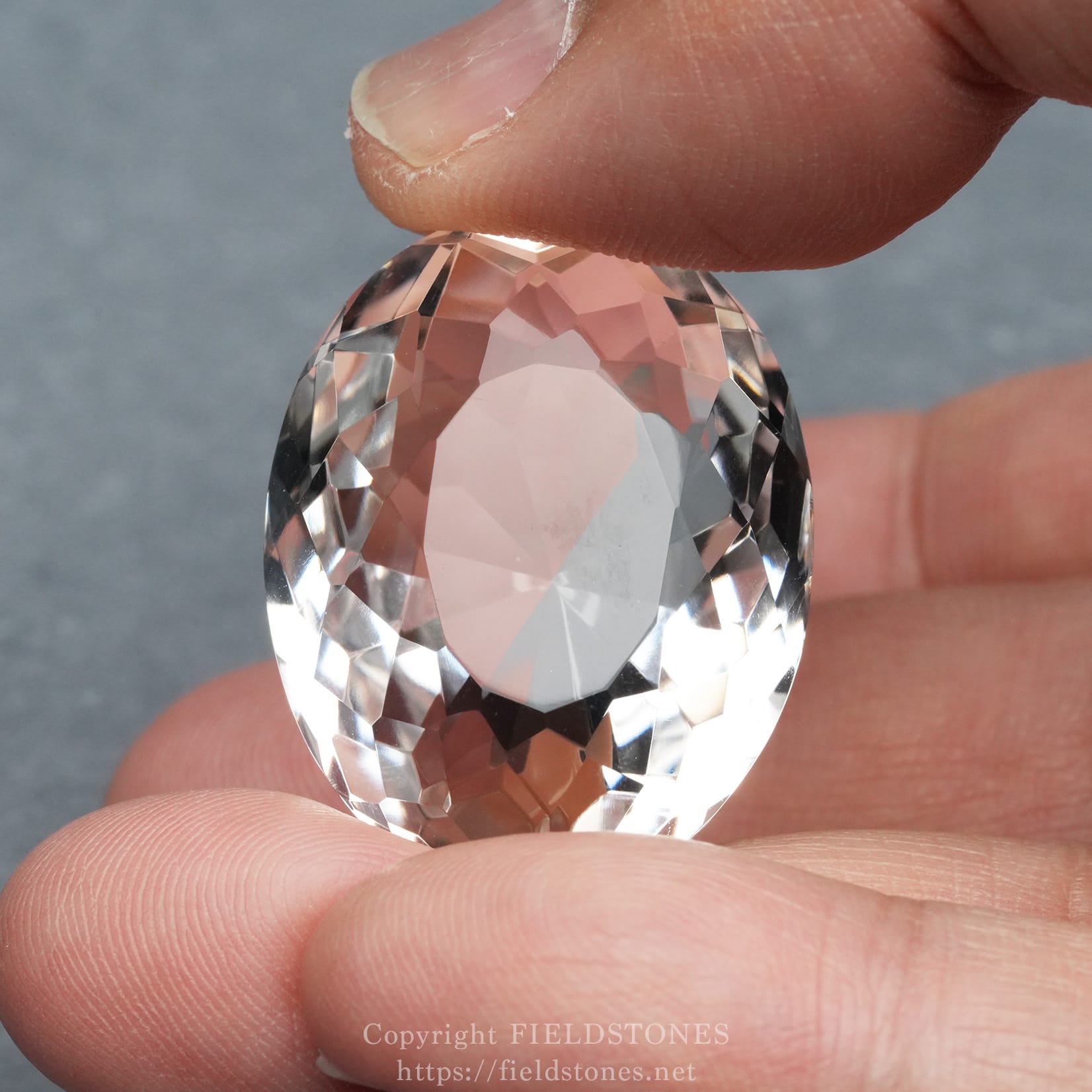 6537-4 - FieldStones Close-up of a faceted pink crystal being held between a thumb and forefinger, showing its many facets and translucence.
