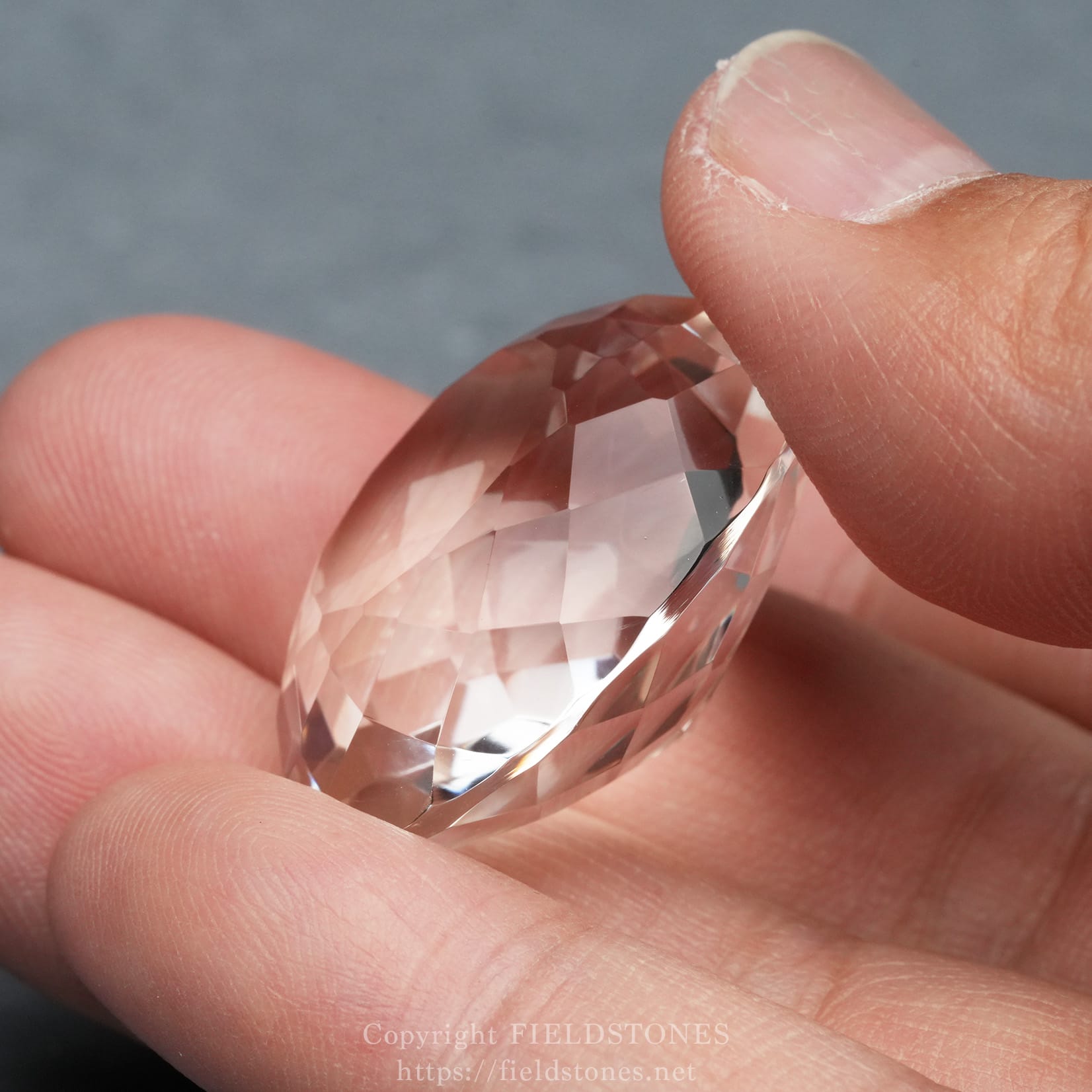 6537-6 - FieldStones Close-up of a faceted transparent gem being held between two fingers, showing its cut and clarity against a soft background.