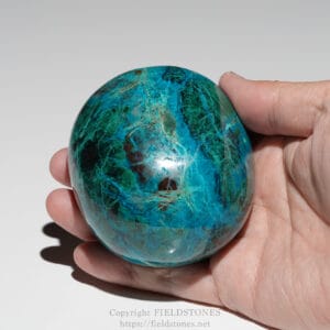 Hand holding a polished blue-green stone sphere (malachite) with natural veining and a glossy finish.