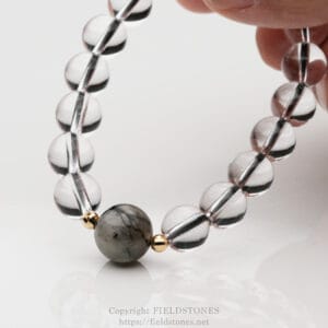 Close-up of a beaded bracelet with clear glass or crystal beads and gold spacers, held by a hand on a reflective surface.