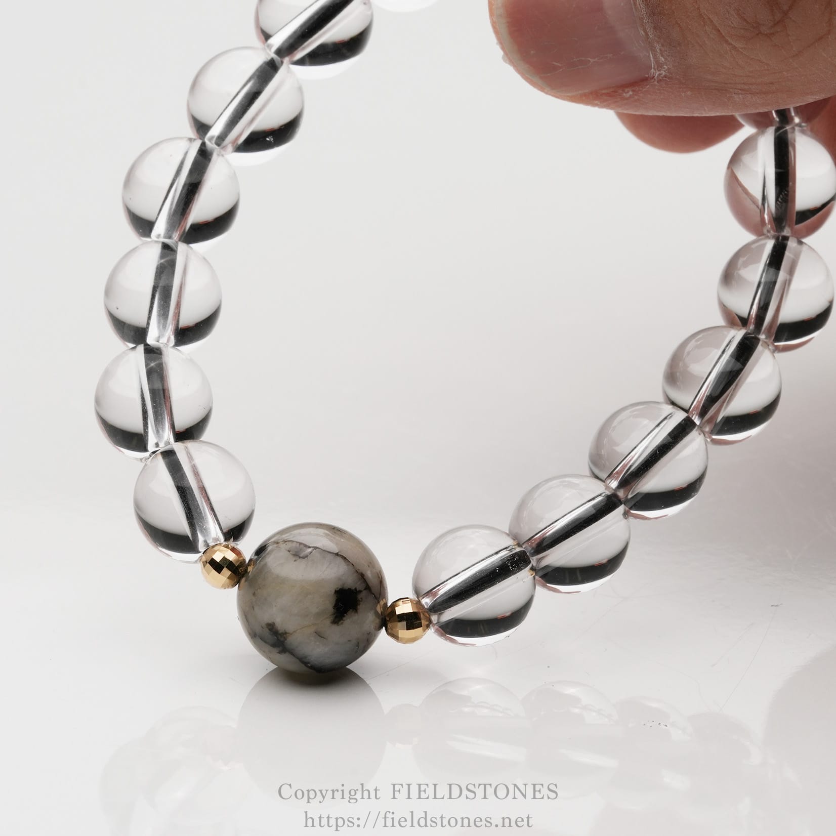 6547-1 - FieldStones Close-up of a beaded bracelet held by a finger, featuring clear glass-like beads and a grey marbled stone bead with small gold spacers on a white reflective surface