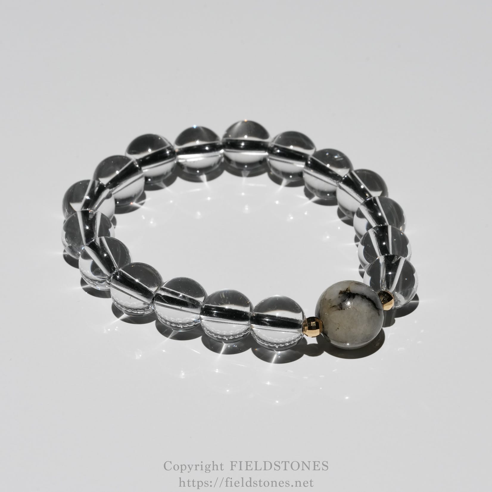 6547-3 - FieldStones Clear glass bead bracelet arranged in a circle on a reflective surface, with a single marbled gray bead and gold spacers.