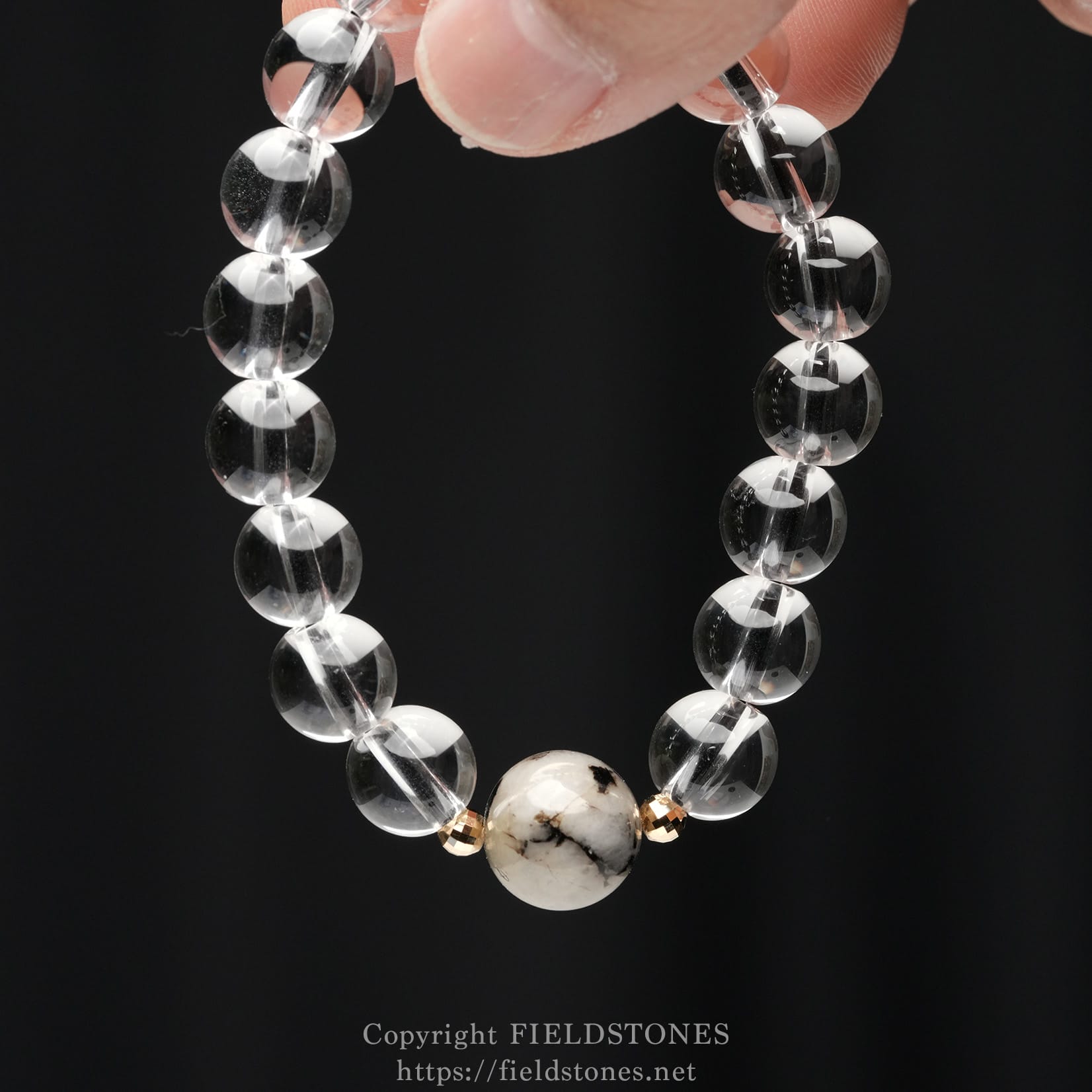 6547-4 - FieldStones Close-up of a beaded bracelet with clear glass or crystal beads and a marbled white central bead, accented by small gold spacers, against a black background.