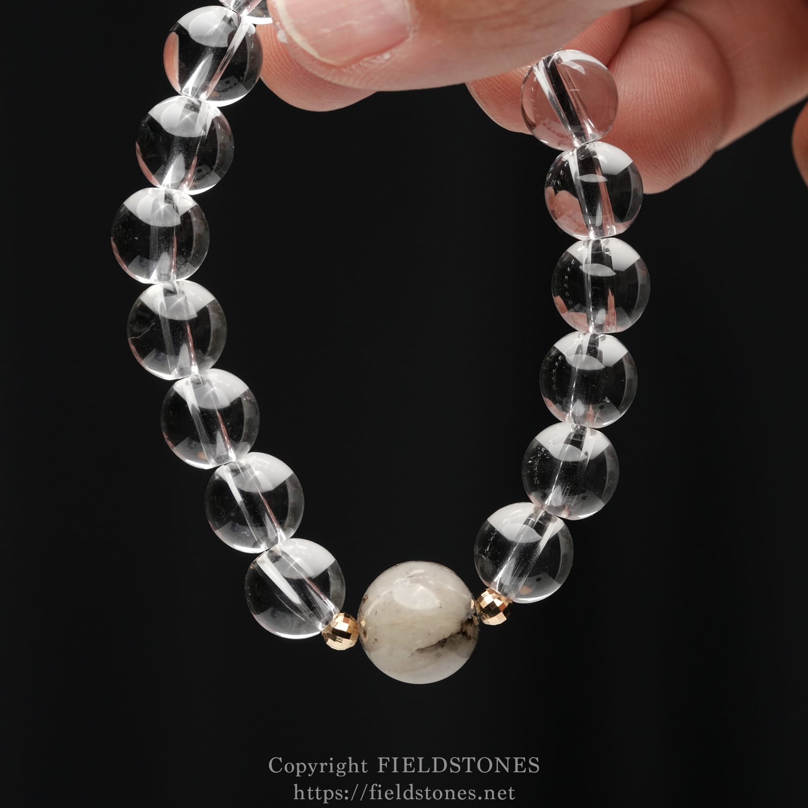 6547-5 - FieldStones Close-up of a beaded bracelet with clear faceted glass beads and a larger pale marbled centerpiece bead, accented by small gold spacers against a black background.