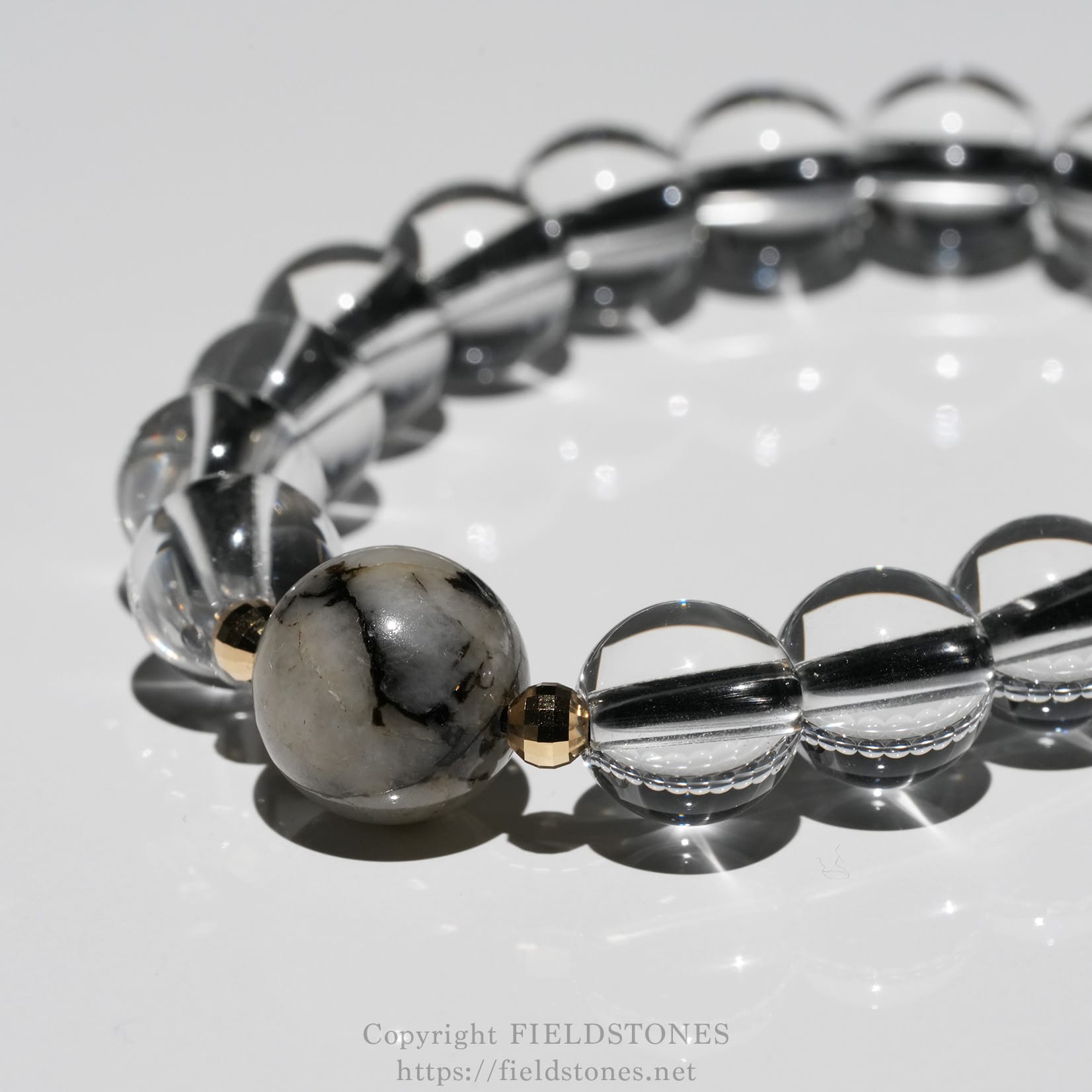 6547-6 - FieldStones Close-up of a beaded bracelet featuring a large marbled gray stone bead, clear glass beads, and a small gold spacer bead on a glossy surface.