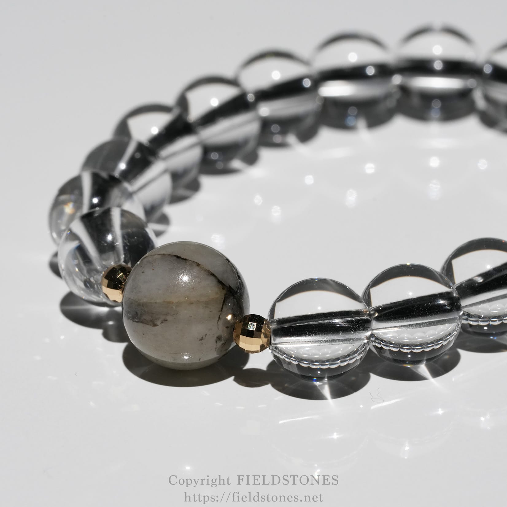 6547-7 - FieldStones Close-up of a beaded bracelet with clear glass beads, small gold spacers, and a marbled gray stone centerpiece on a white surface.