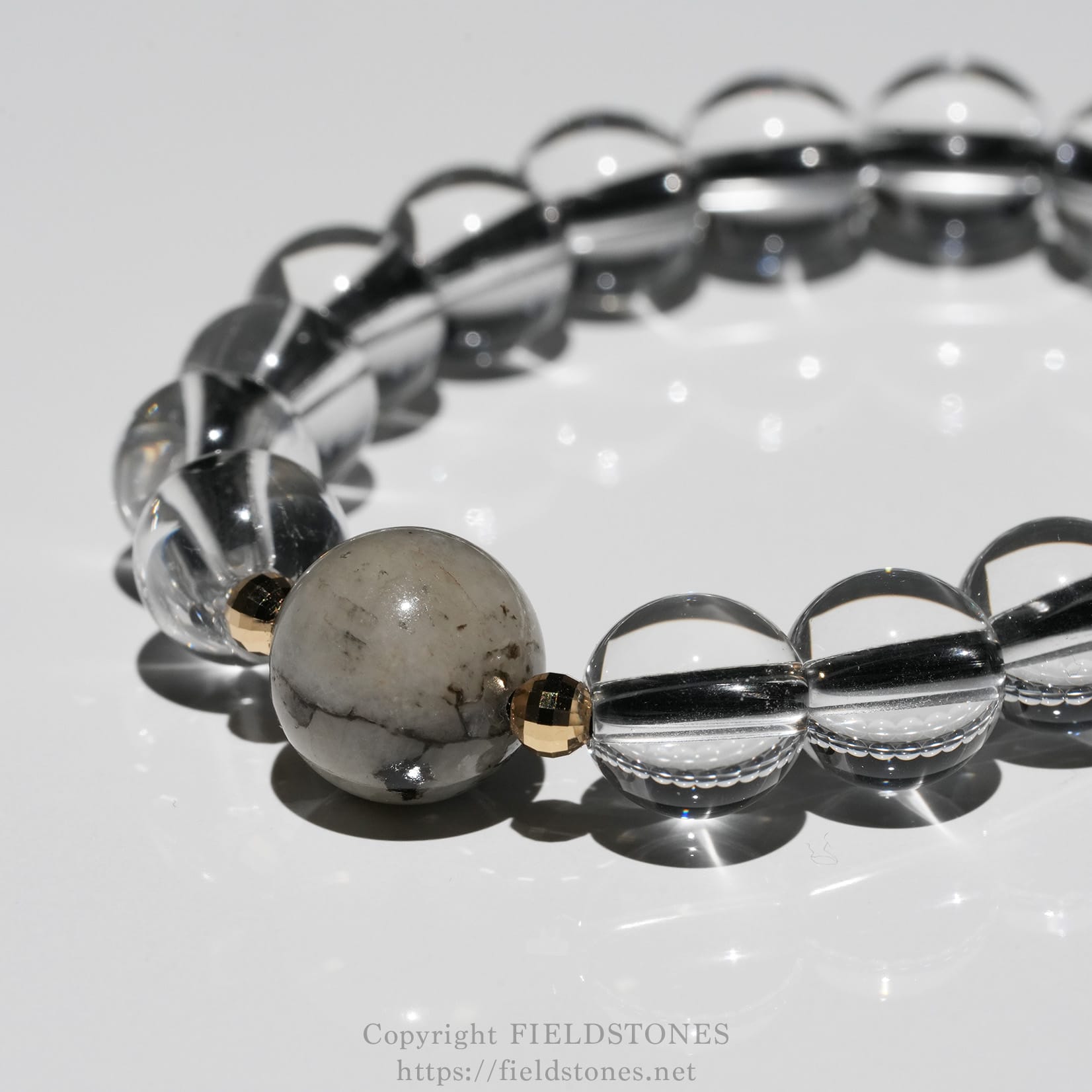 6547-8 - FieldStones Close-up of a beaded bracelet with clear glass beads, a marbled gray stone bead, and small gold spacers on a white background.