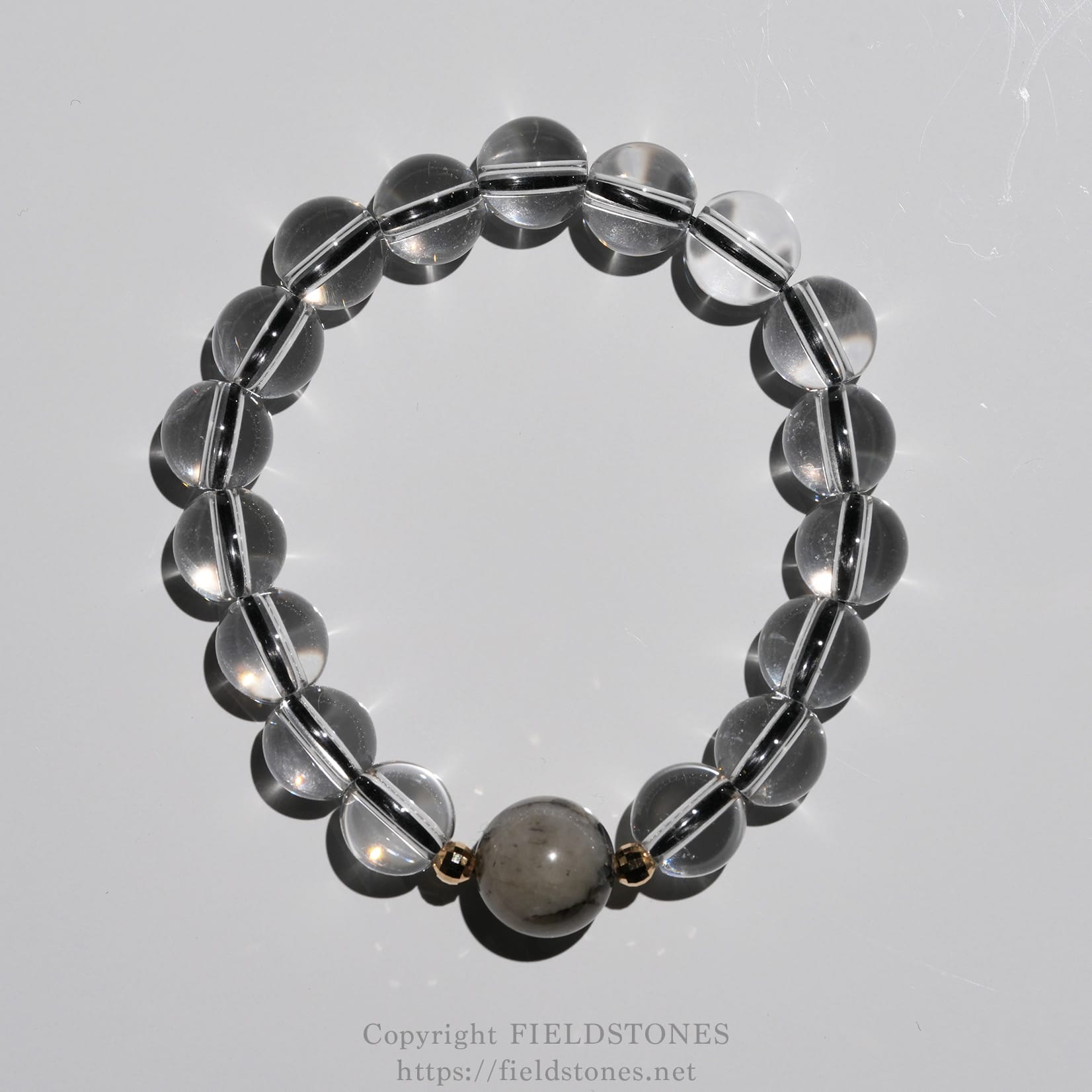 6547-9 - FieldStones Bracelet made of clear glass beads on a reflective surface, with a larger marbled central bead and gold spacers nearby