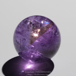 Polished purple amethyst sphere resting on a reflective surface, showing internal fractures and bright highlights.