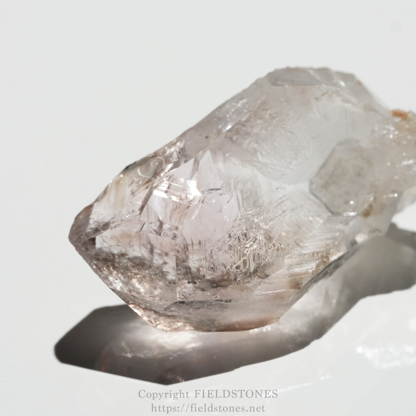 6562-2 - FieldStones Close-up of a rough, pale pinkish quartz crystal on a white surface, showing sharp facets and a soft shadow beneath it.