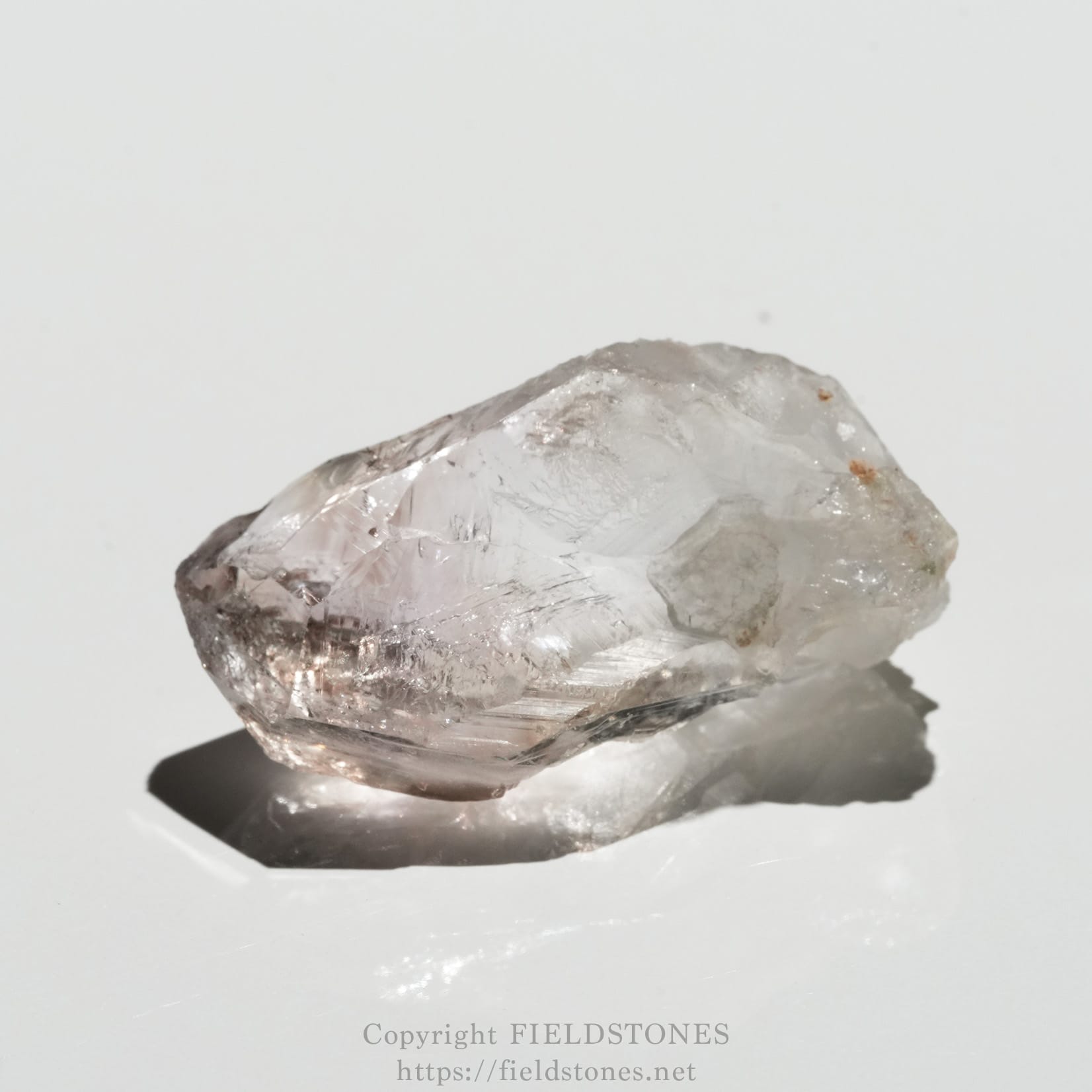 6562-4 - FieldStones Close-up of a clear quartz crystal resting on a white surface, casting a shadow beneath it.