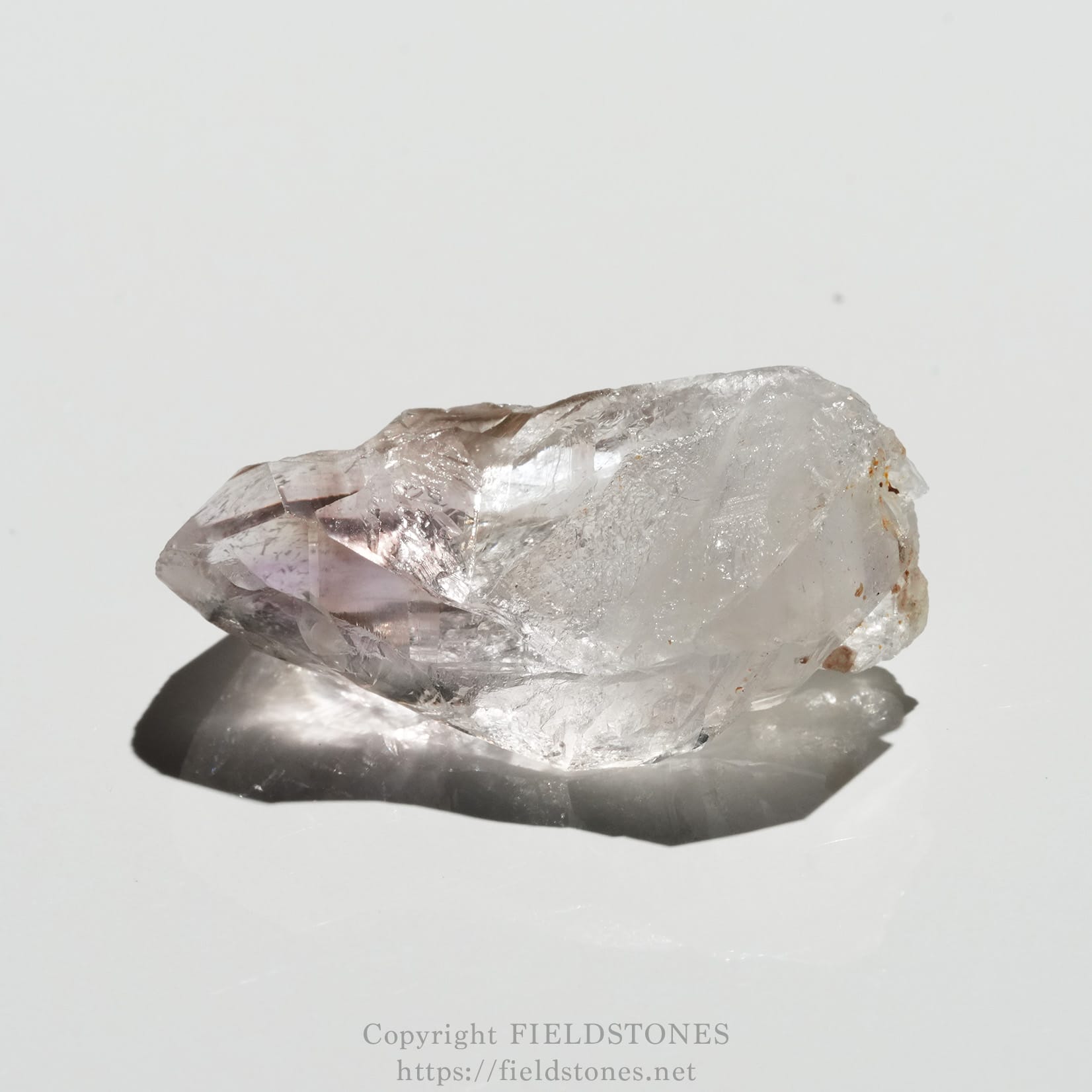 6562-5 - FieldStones Clear quartz crystal resting on a white surface with soft reflections and a faint pink tint near the facets.