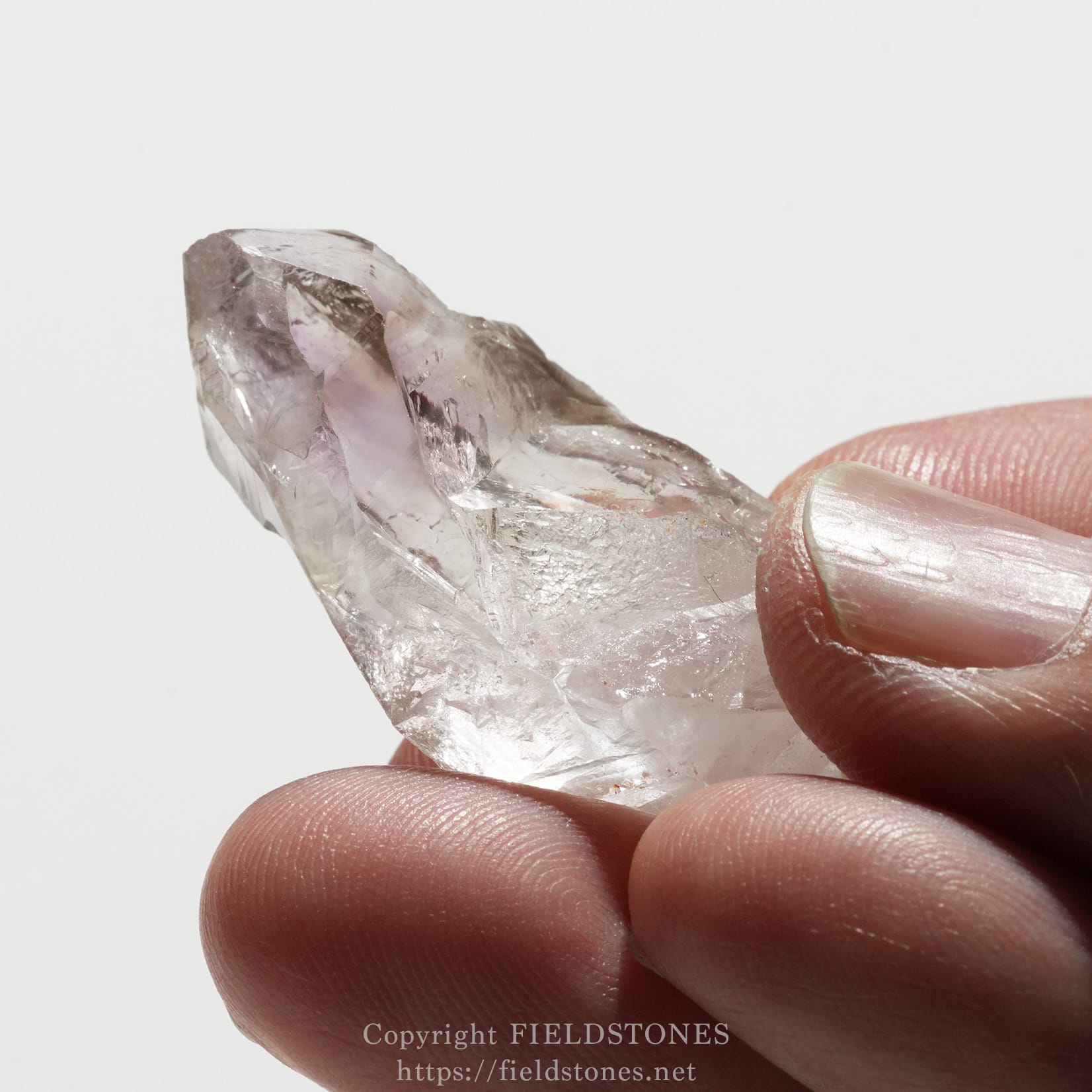 6562-8 - FieldStones Close-up of a translucent quartz crystal being held between the thumb and forefinger, pale and cloudy with sharp facets.