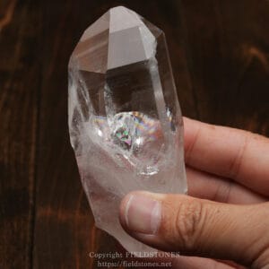 Close-up of a clear quartz crystal being held between two fingers against a dark wooden background.