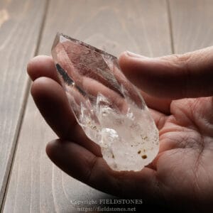 Person holding a large clear quartz crystal between fingertips over a wooden surface.