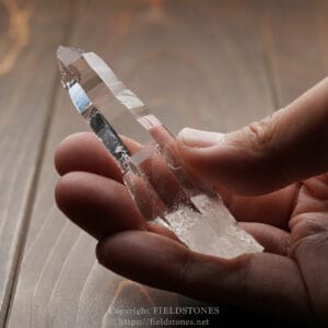 Clear quartz crystal point held between thumb and forefinger over a wooden surface.