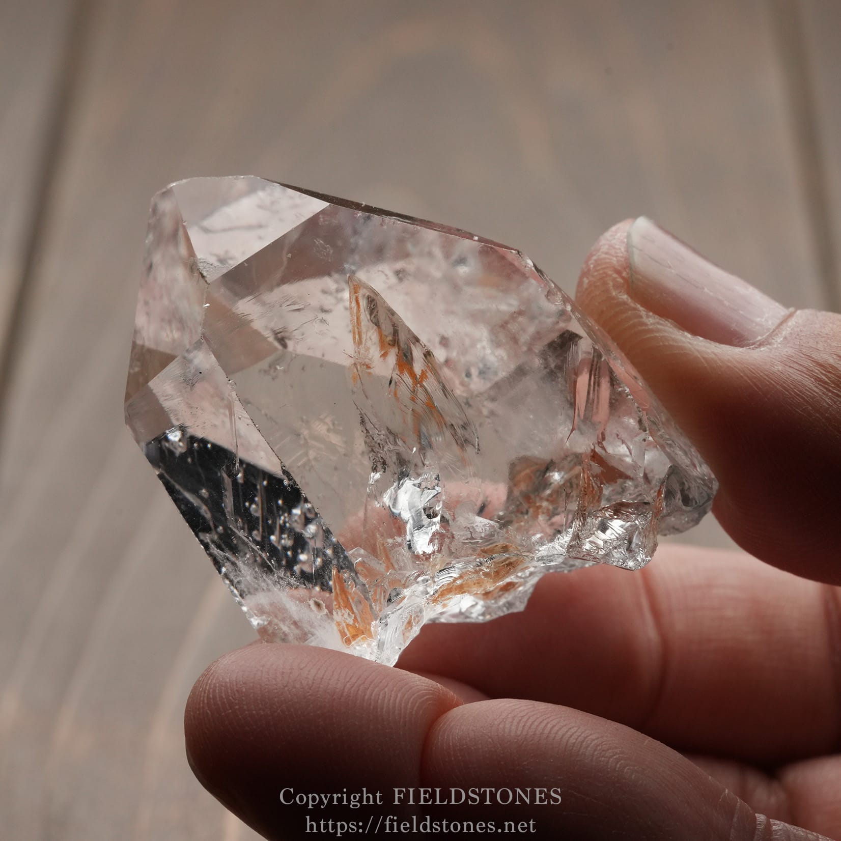 6571-11 - FieldStones Close-up of a large, translucent quartz crystal held between thumb and index finger, showing sharp facets and clarity between the fingers.
