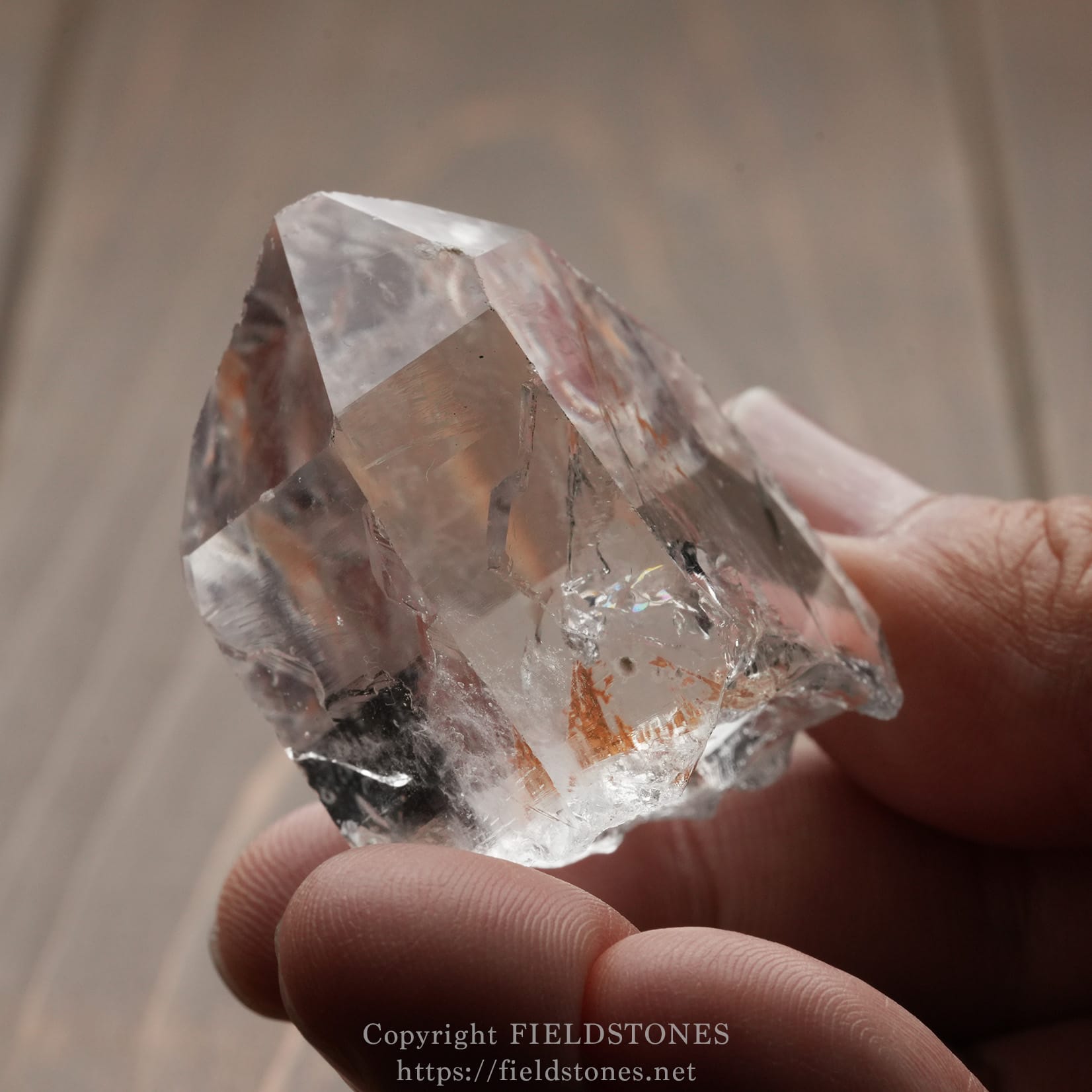 6571-12 - FieldStones Clear quartz crystal held between fingers, showing sharp facets and natural inclusions when light passes through it, close-up.