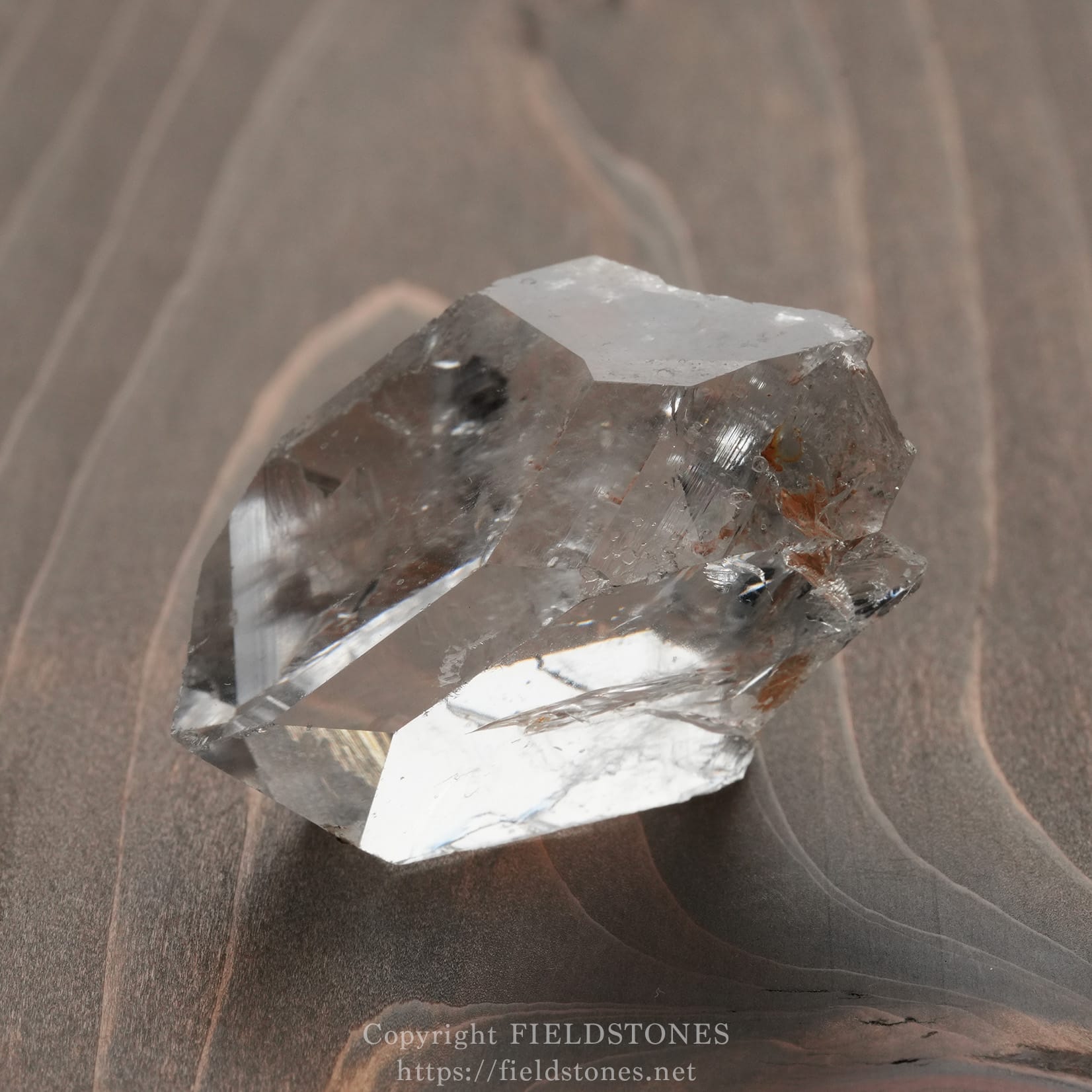 6571-13 - FieldStones Clear quartz crystal with sharp facets resting on a dark wooden surface.