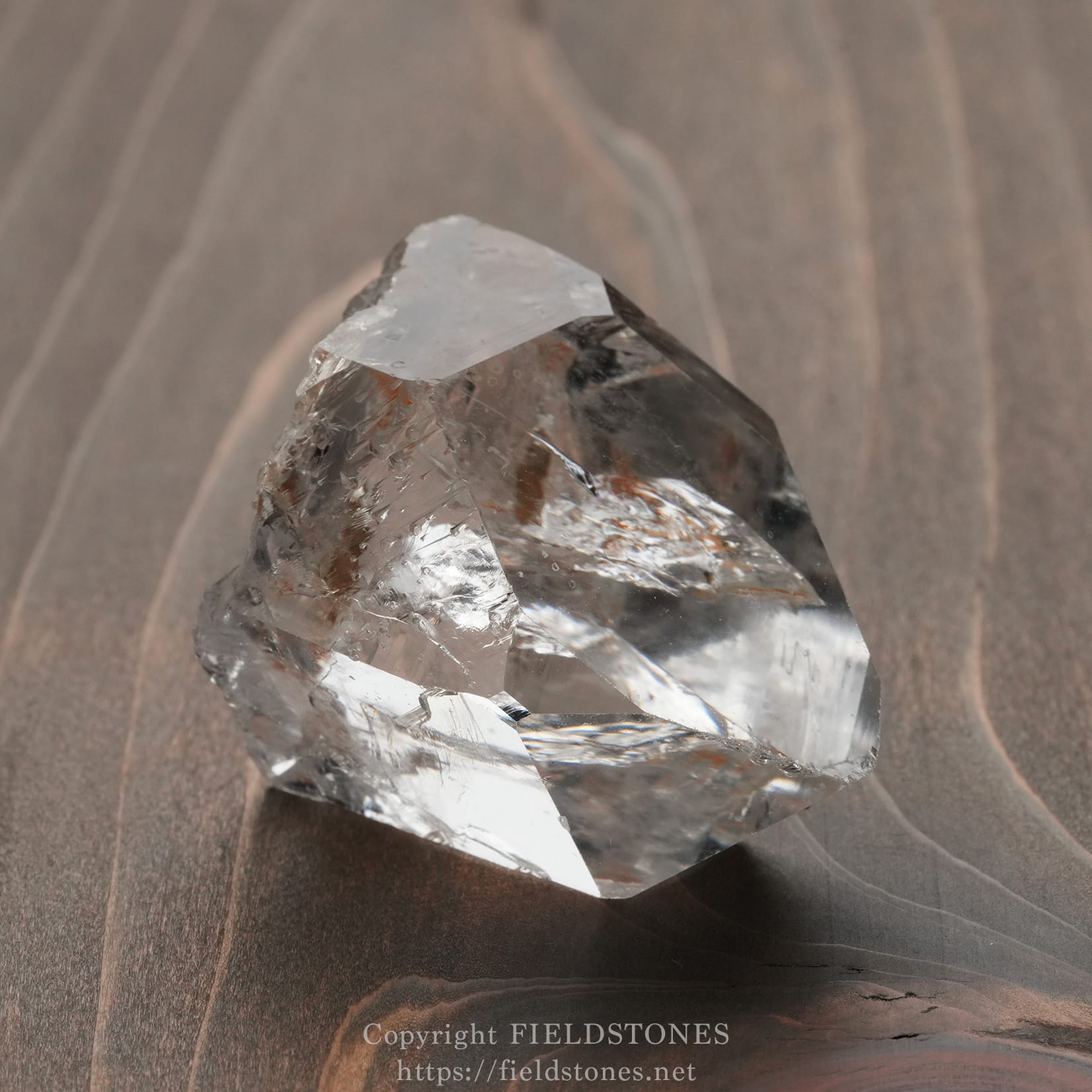 6571-14 - FieldStones Close-up of a clear quartz crystal resting on a wooden surface, showing sharp facets and reflections on the grainy background.