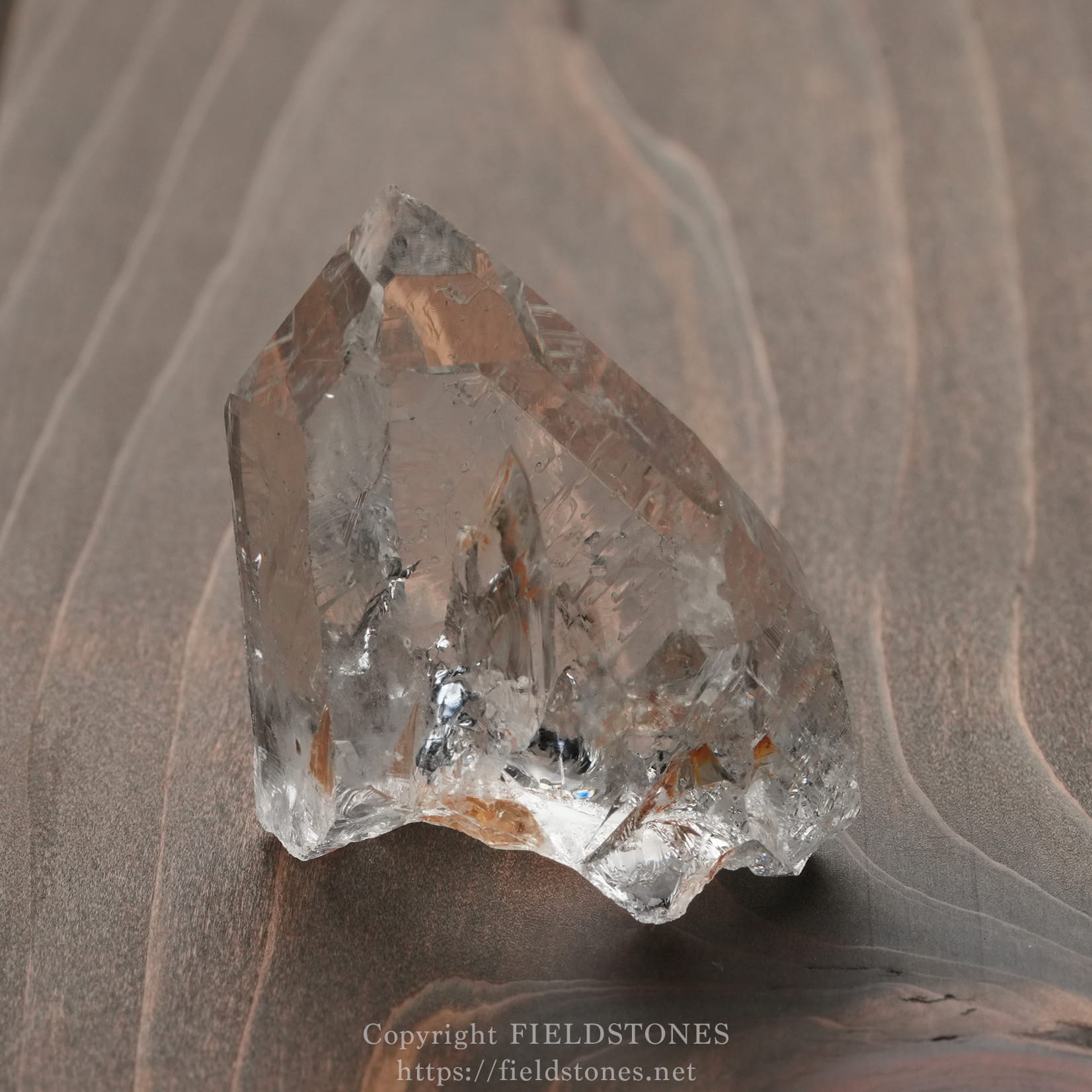 6571-15 - FieldStones Clear quartz crystal standing upright on a brown wooden surface, showing sharp facets and translucence.