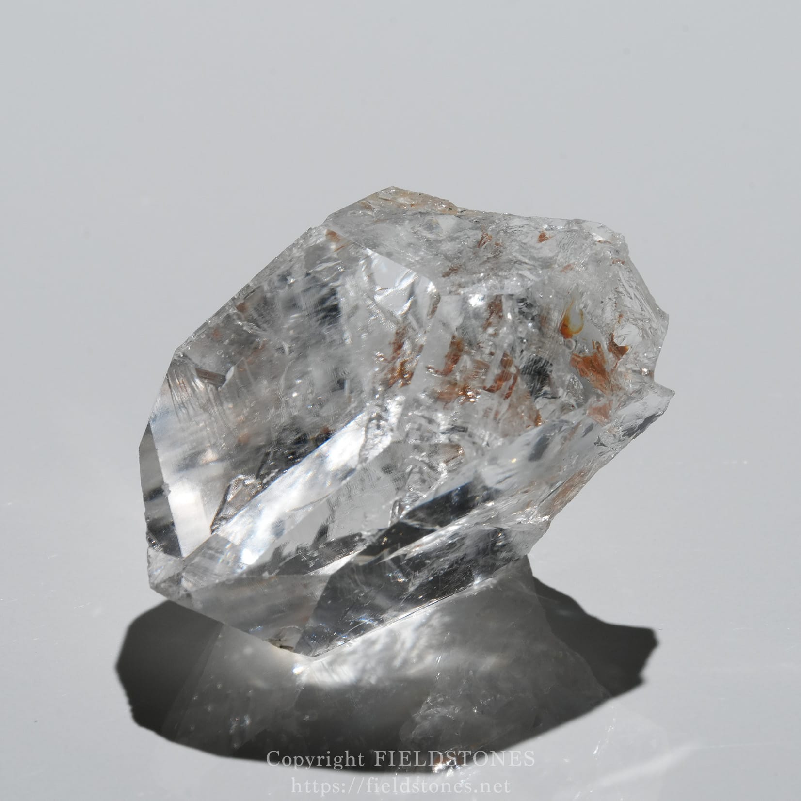 6571-3 - FieldStones Close-up of a clear quartz/crystal gemstone with sharp facets on a pale gray background, casting a dark shadow.