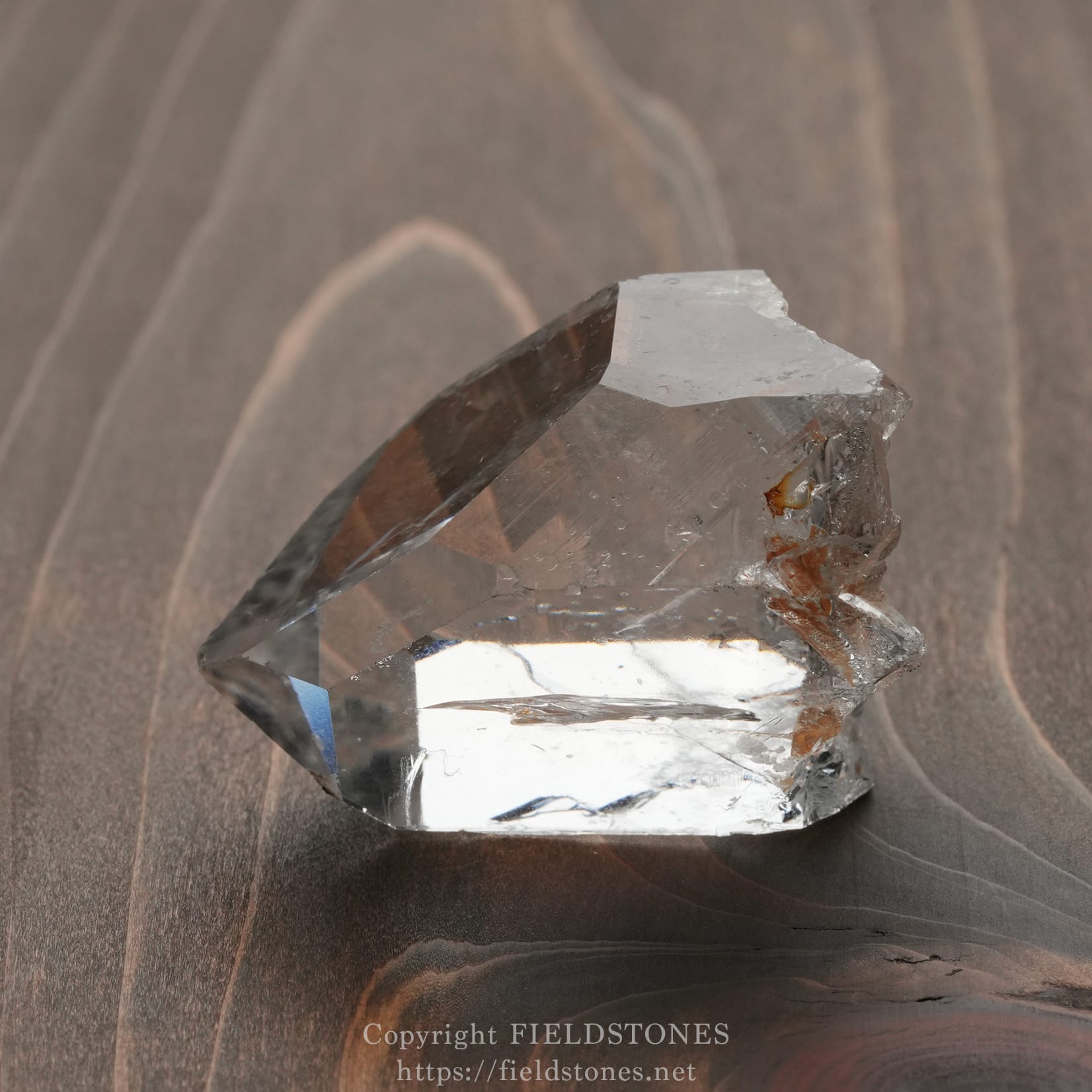 6571-5 - FieldStones Clear quartz crystal with sharp facets resting on a dark wooden surface.