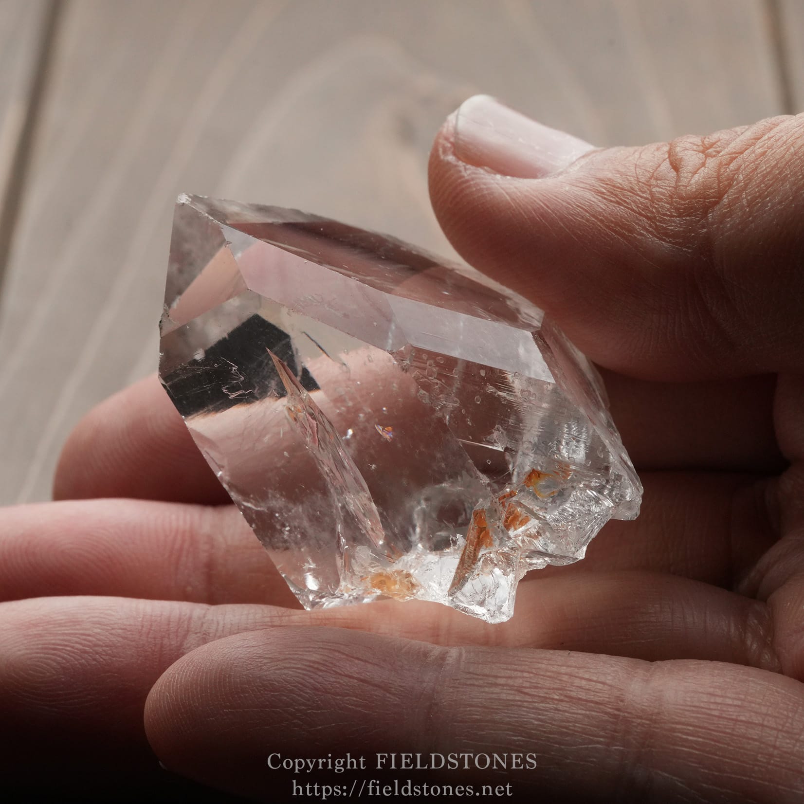 6571-7 - FieldStones Close-up of a clear quartz crystal held between fingertips over a neutral background, showing its translucent facets and small internal flaws.
