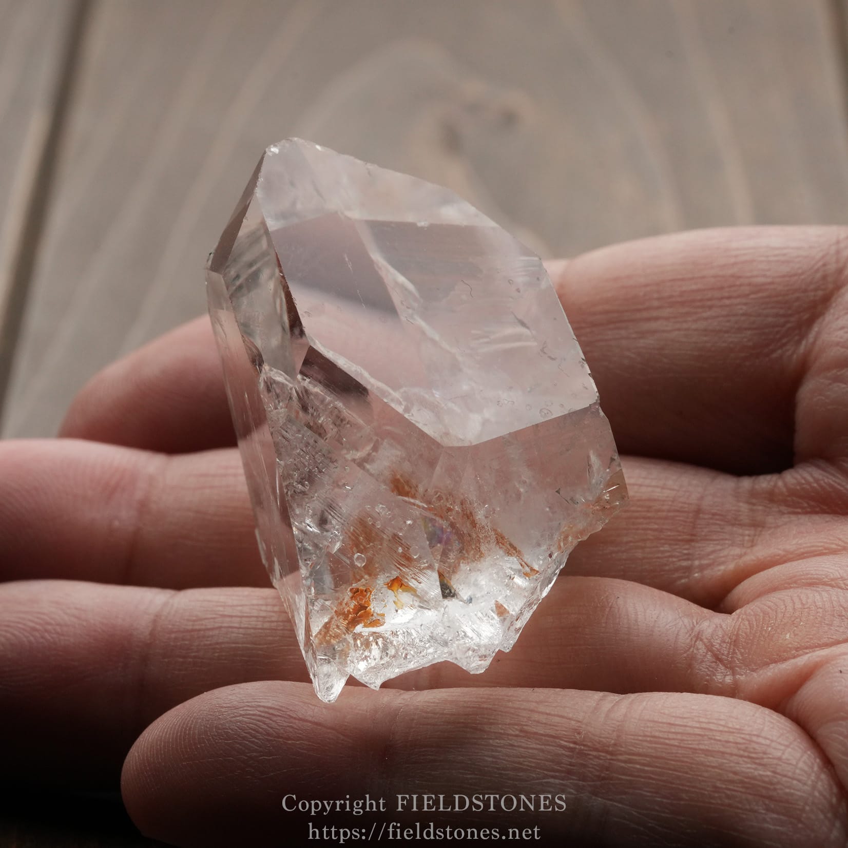 6571-9 - FieldStones Clear quartz crystal held between fingers, with orange mineral inclusions, against a wooden background.