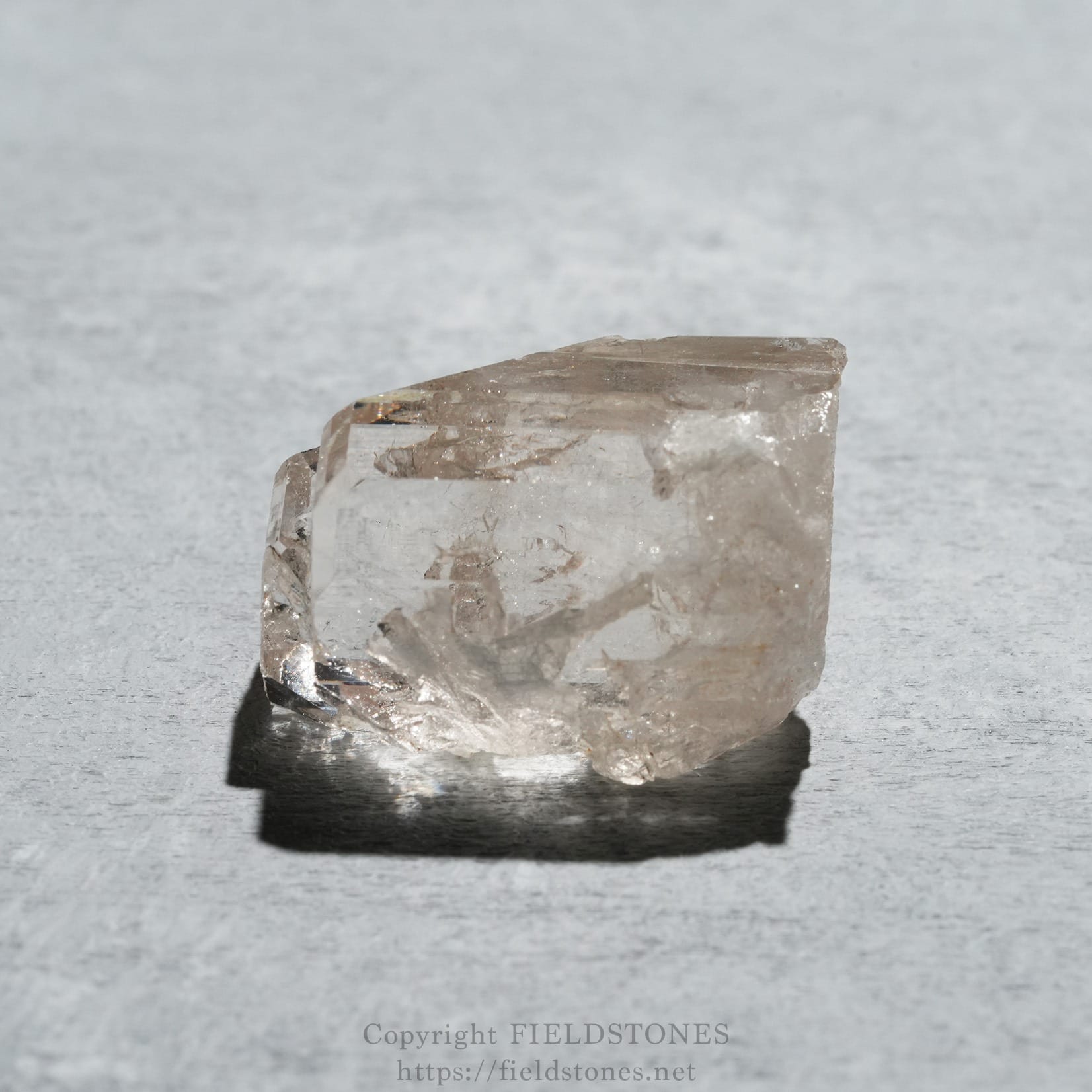 6576-2 - FieldStones Transparent quartz crystal shard resting on a light gray surface, showing its faceted edges and subtle reflections