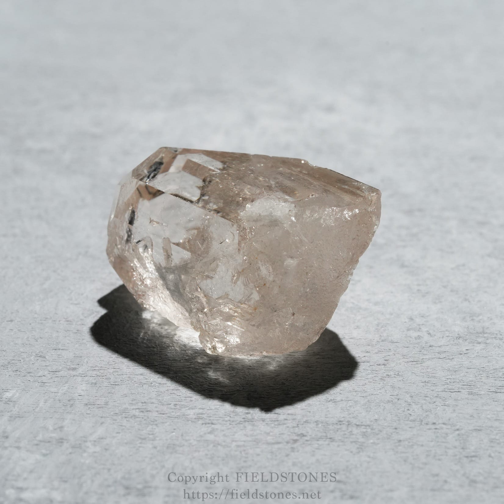 6576-3 - FieldStones Clear quartz crystal on a gray surface with a soft shadow beneath it, showing faceted faces and a pale pink-tawn color.
