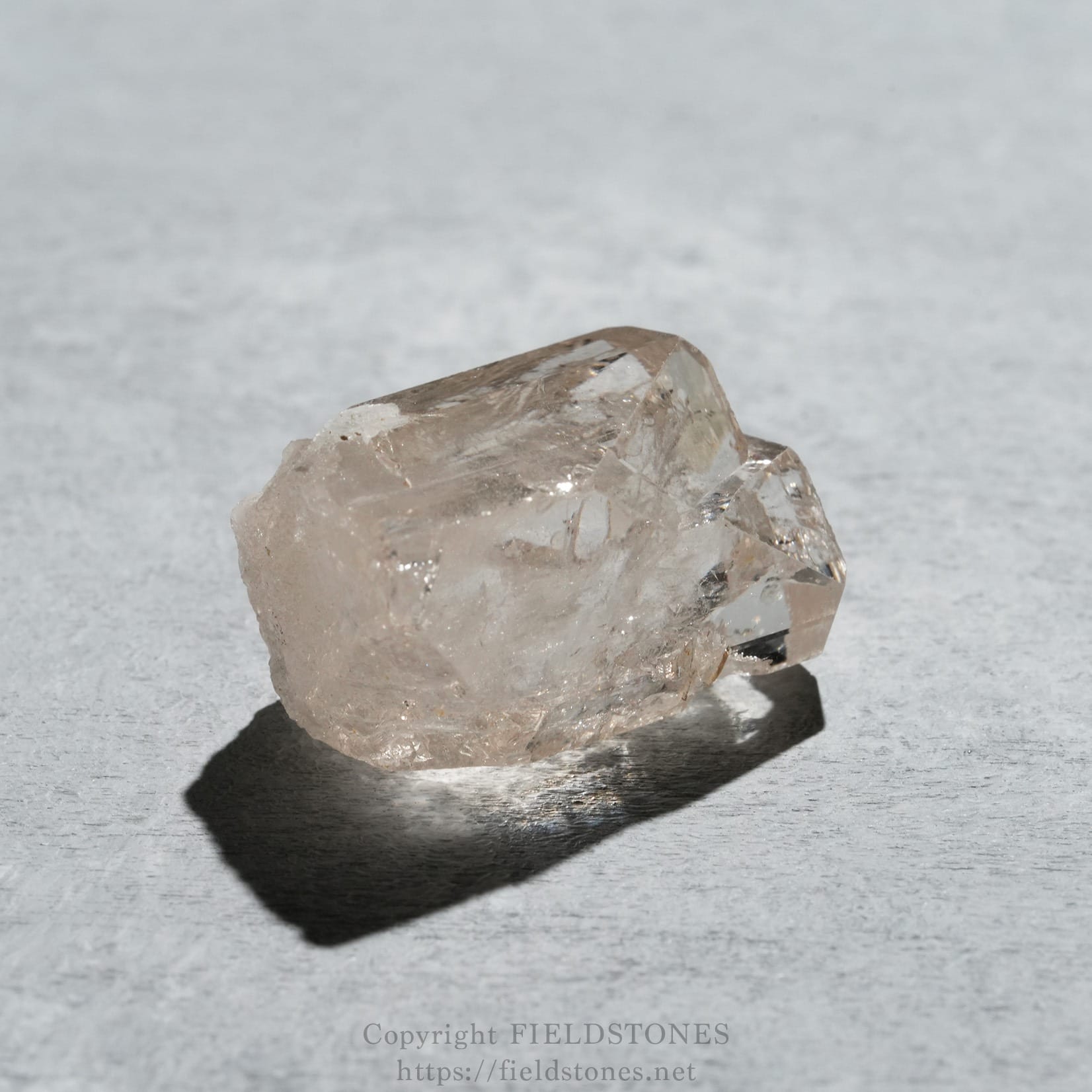 6576-4 - FieldStones A rough, translucent quartz crystal on a gray textured surface with a dark shadow beneath it, photographed close-up.