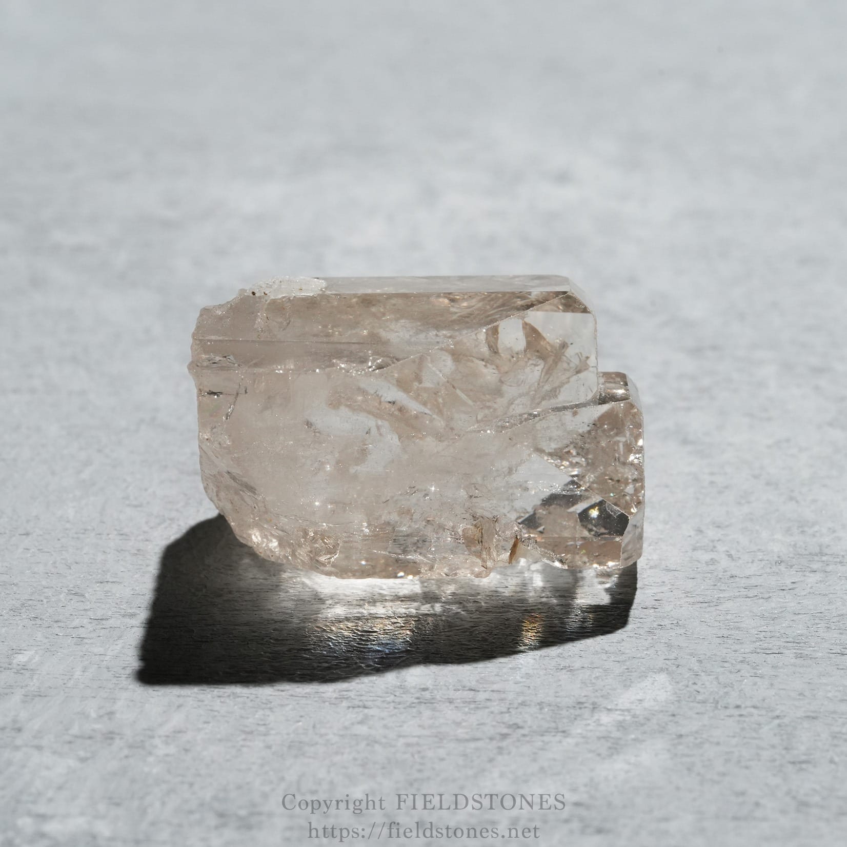 6576-5 - FieldStones Clear quartz crystal resting on a textured gray surface, with a dark shadow beneath it.