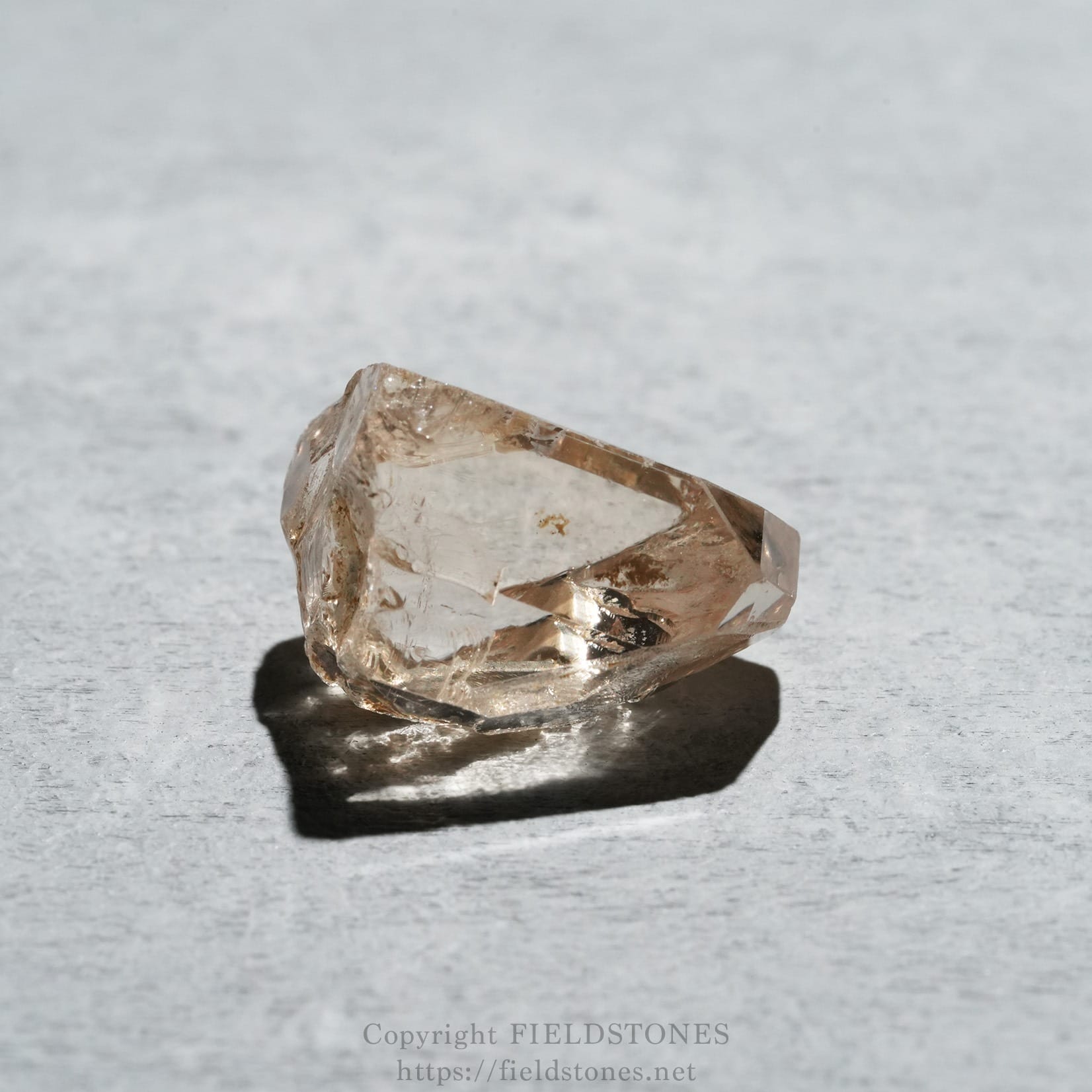 6578-1 - FieldStones Translucent pinkish crystal resting on a light gray textured surface with a shadow beneath it