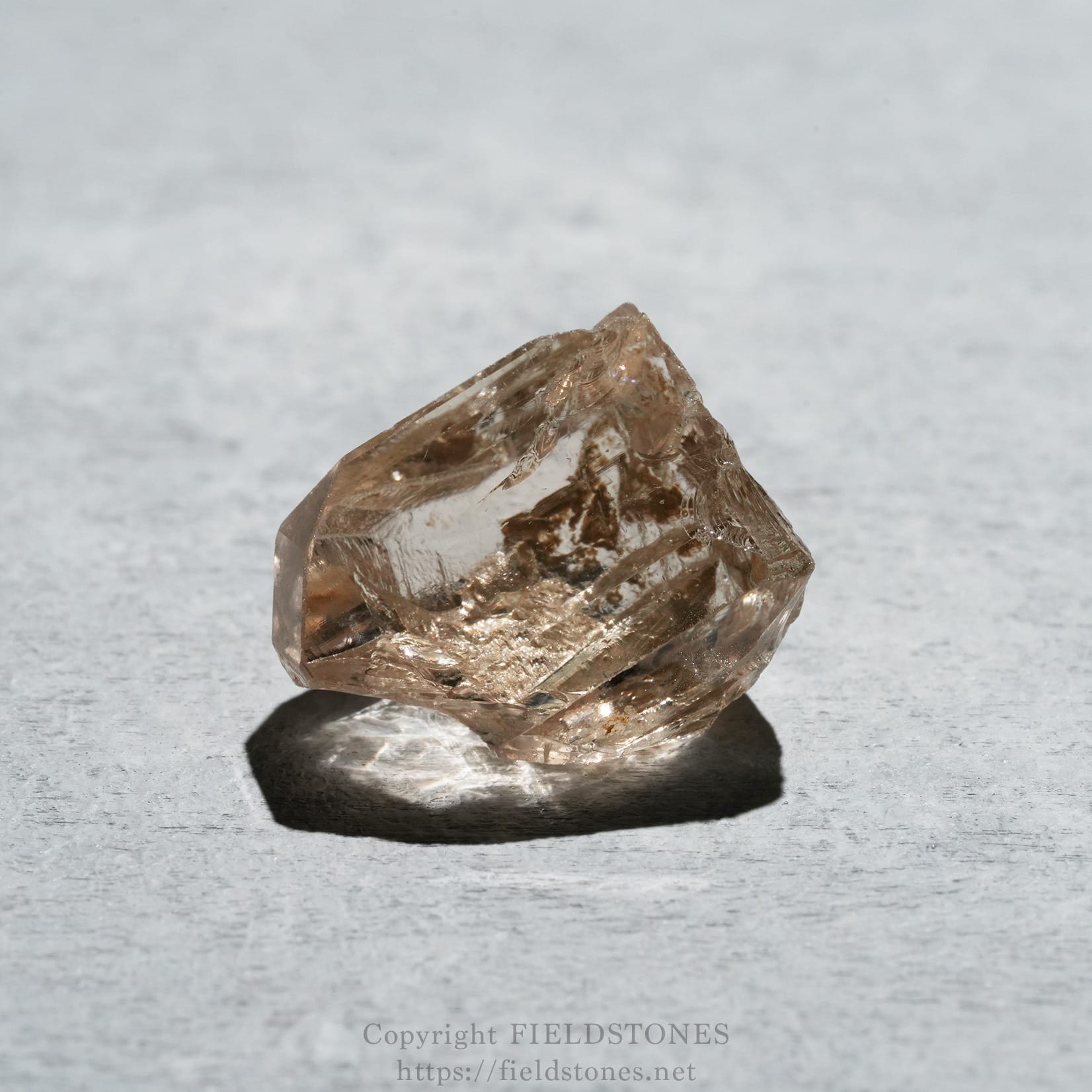 6578-3 - FieldStones Pinkish-brown crystal resting on a light grey textured surface, casting a dark shadow beneath it.