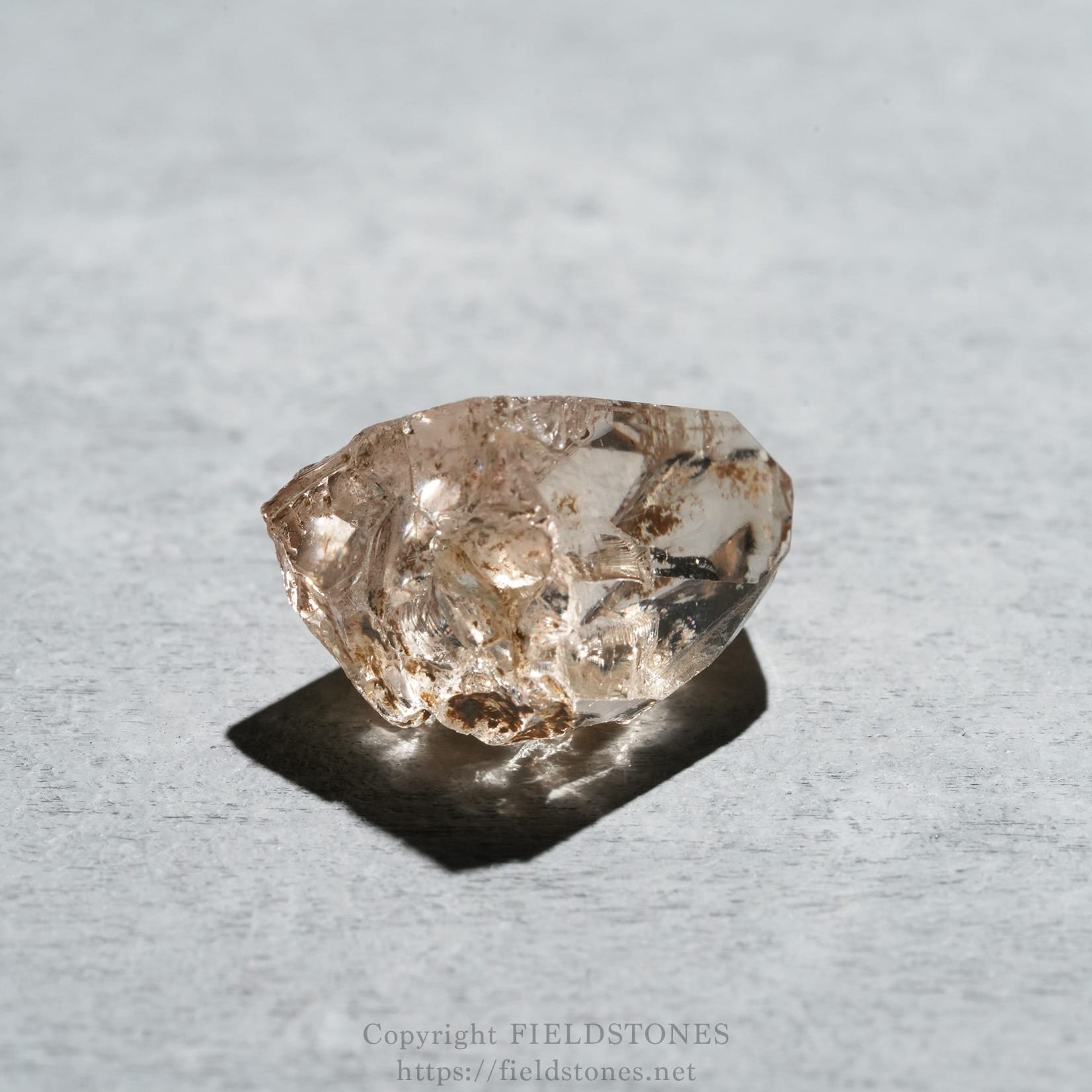 6578-6 - FieldStones Rough, translucent crystal (quartz) resting on a light gray textured surface, photographed up close.