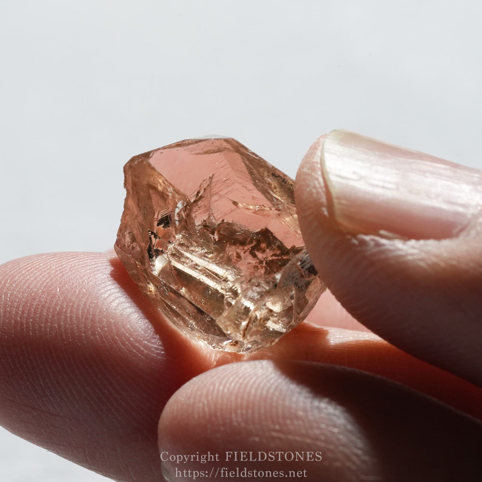 6578-8 - FieldStones Close-up of a pink crystal held between two fingers against a pale background, showing its facets and translucence.
