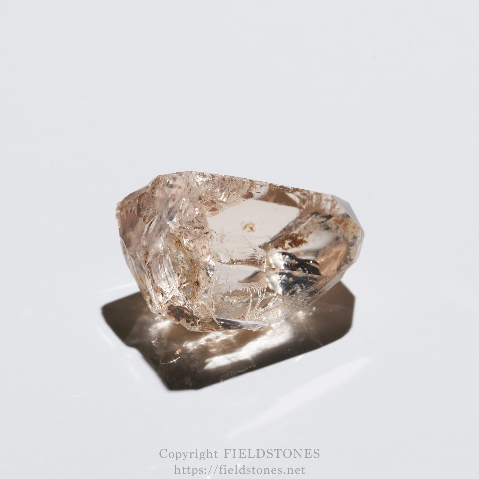6578-9 - FieldStones Rough pinkish crystal or gemstone resting on a white surface, casting a shadow.
