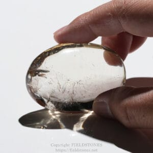 Close-up of a hand holding a polished, transparent quartz palm stone against a white background.