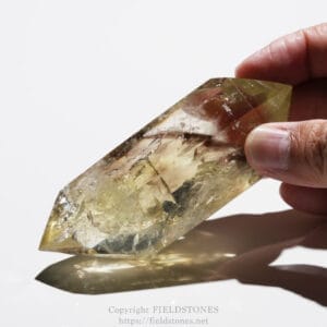 Close-up of a hand holding a large, faceted quartz crystal point, sunlight reflecting off its translucent yellow-brown facets.
