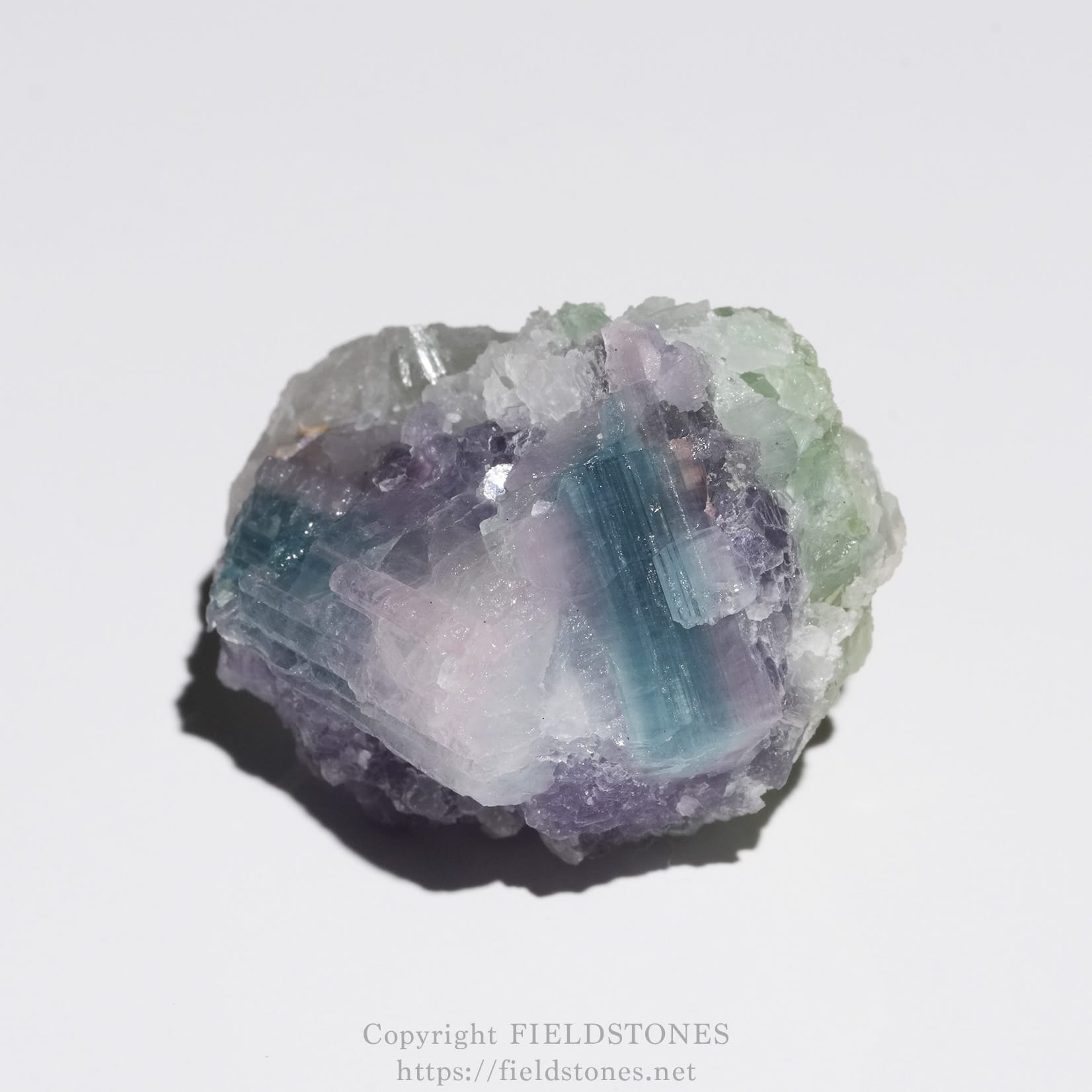 6645-3 - FieldStones Close-up of a rough, multi-hued crystal cluster with purple, blue, and pale green facets on a white background. Watermark text is visible at the bottom.