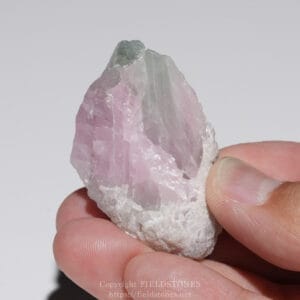 Close-up of a rough pink quartz crystal held between fingers, with a small green mineral at the top (rose quartz with a green cap).