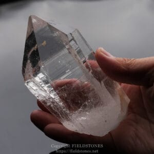 Close-up of a large clear quartz crystal held between a thumb and forefinger, showing sharp facets and subtle inclusions.
