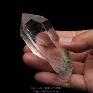 Clear, hexagonal quartz crystal held between fingers against a black background, showing transparency and natural facets.