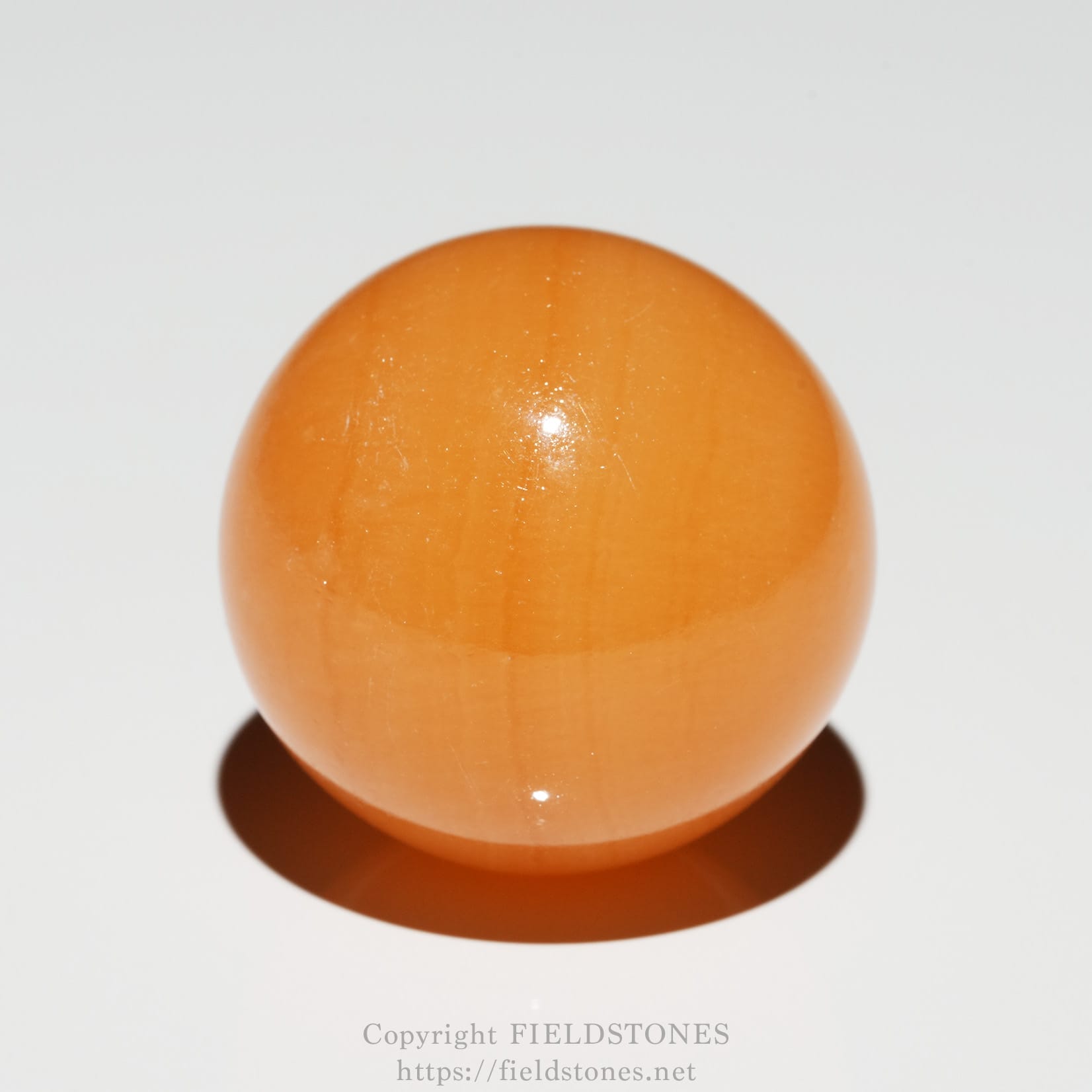 6676-2 - FieldStones Close-up of an orange spherical ball on a white surface with a soft shadow beneath it and a faint watermark at the bottom edge.