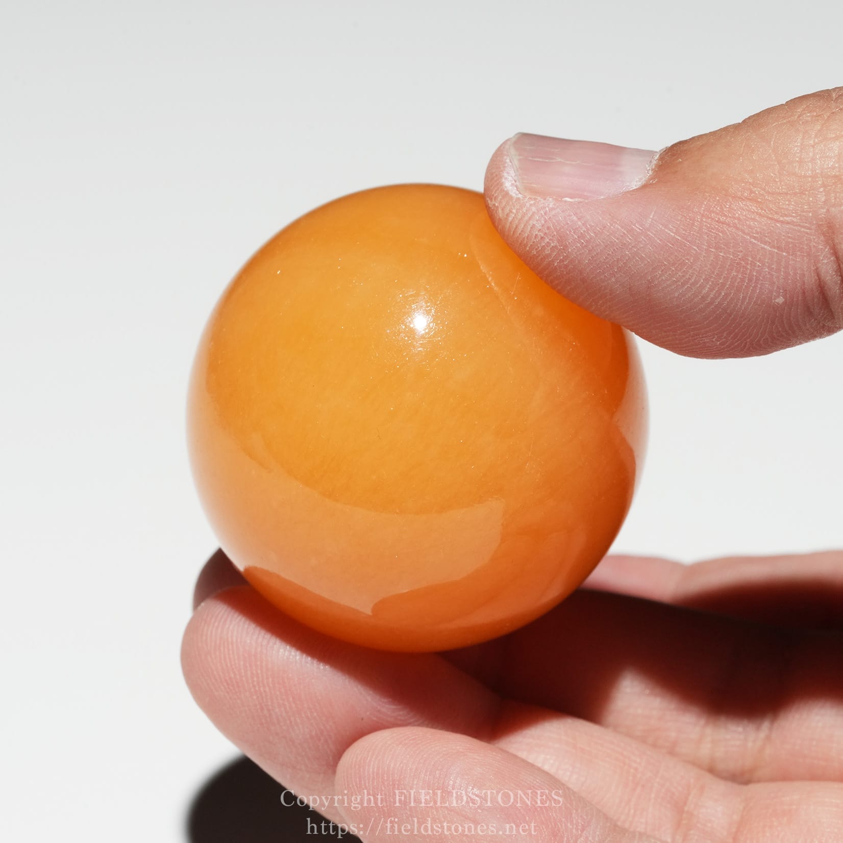 6676-5 - FieldStones Close-up of a thumb and finger pinching a glossy orange ball against a light background.