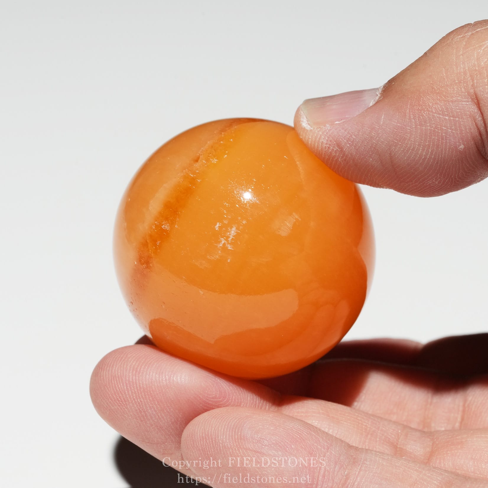 6678-5 - FieldStones Close-up of an orange marble being pinched between two fingers at the edge of the frame, white background.