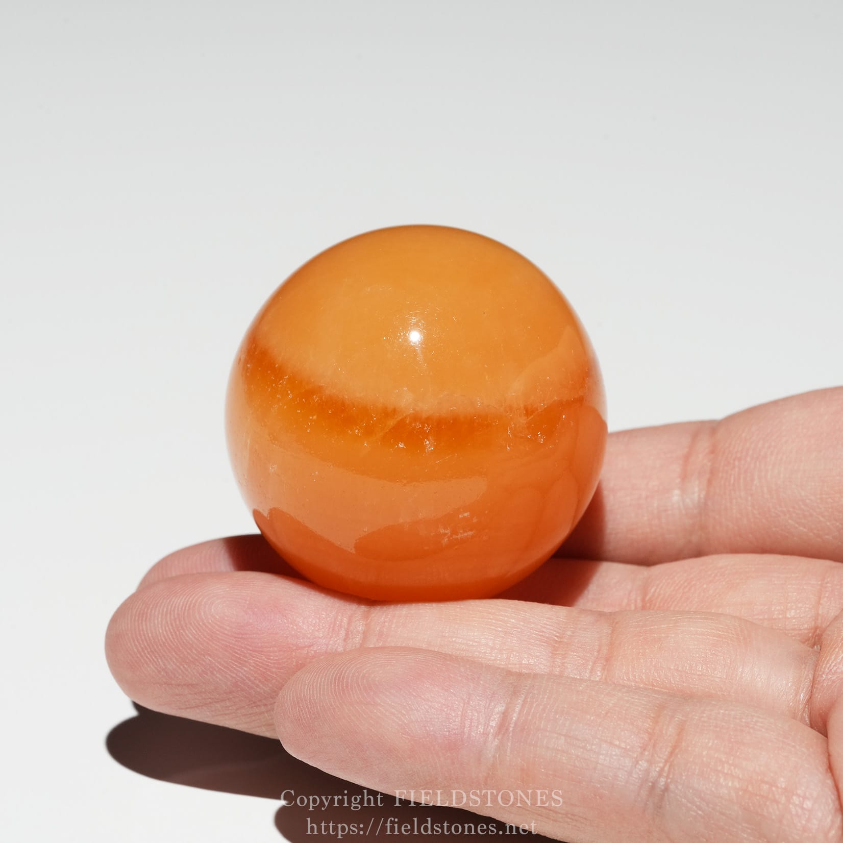 6678-7 - FieldStones Close-up of a smooth orange stone egg held between two fingers, with a glossy surface and subtle banding visible near the bottom edge of the egg.