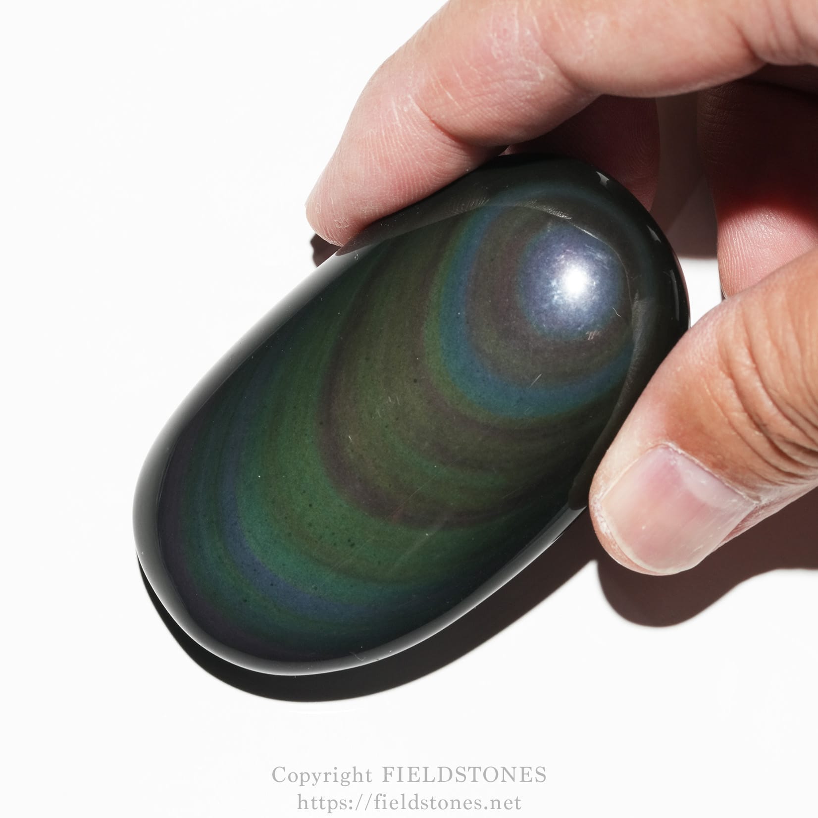 6682-1 - FieldStones Close-up of a dark green, teardrop-shaped polished gemstone with iridescent blue-green swirls, held between a thumb and forefinger.